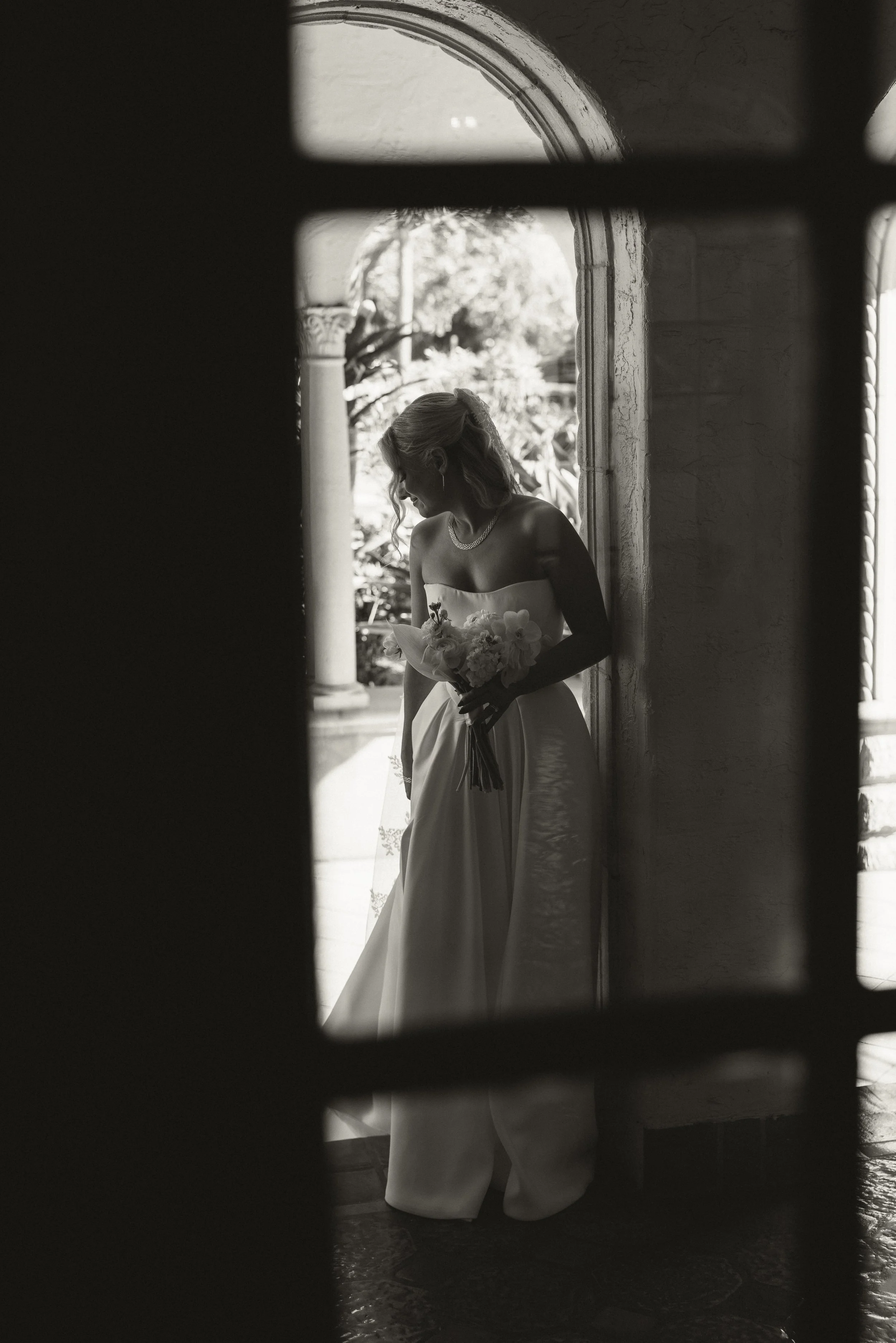 A bride in a wedding dress holding a bouquet of flowers, standing in a doorway, captured in black and white.