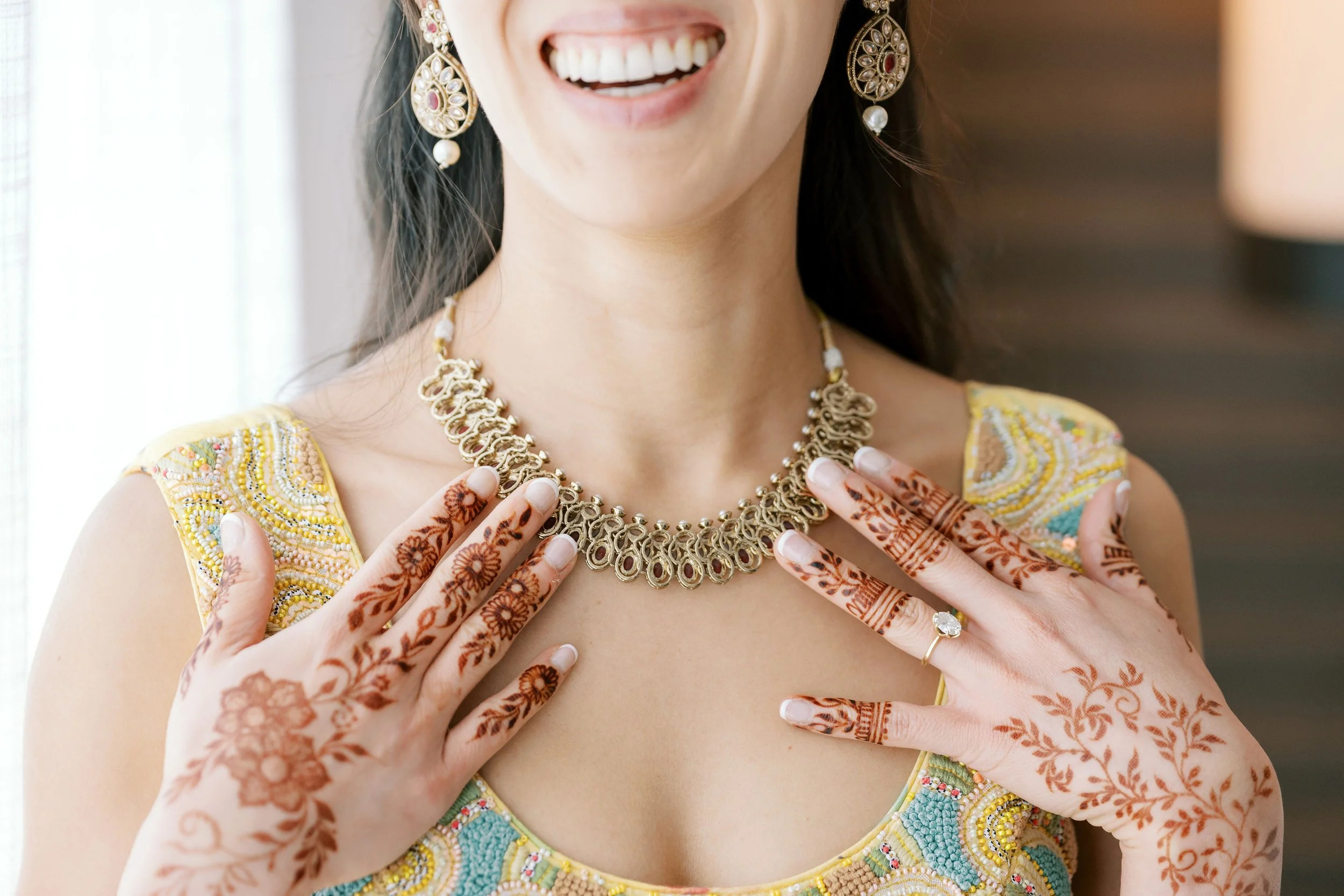 A woman wearing traditional Indian jewelry, including earrings and a necklace, with intricate henna designs on her hands, smiling and adjusting her necklace.