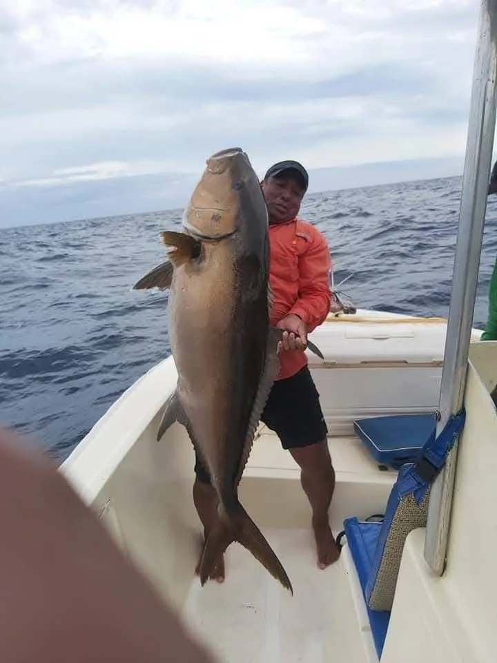 Man in orange jacket and black shorts holding a large fish on a boat at sea.