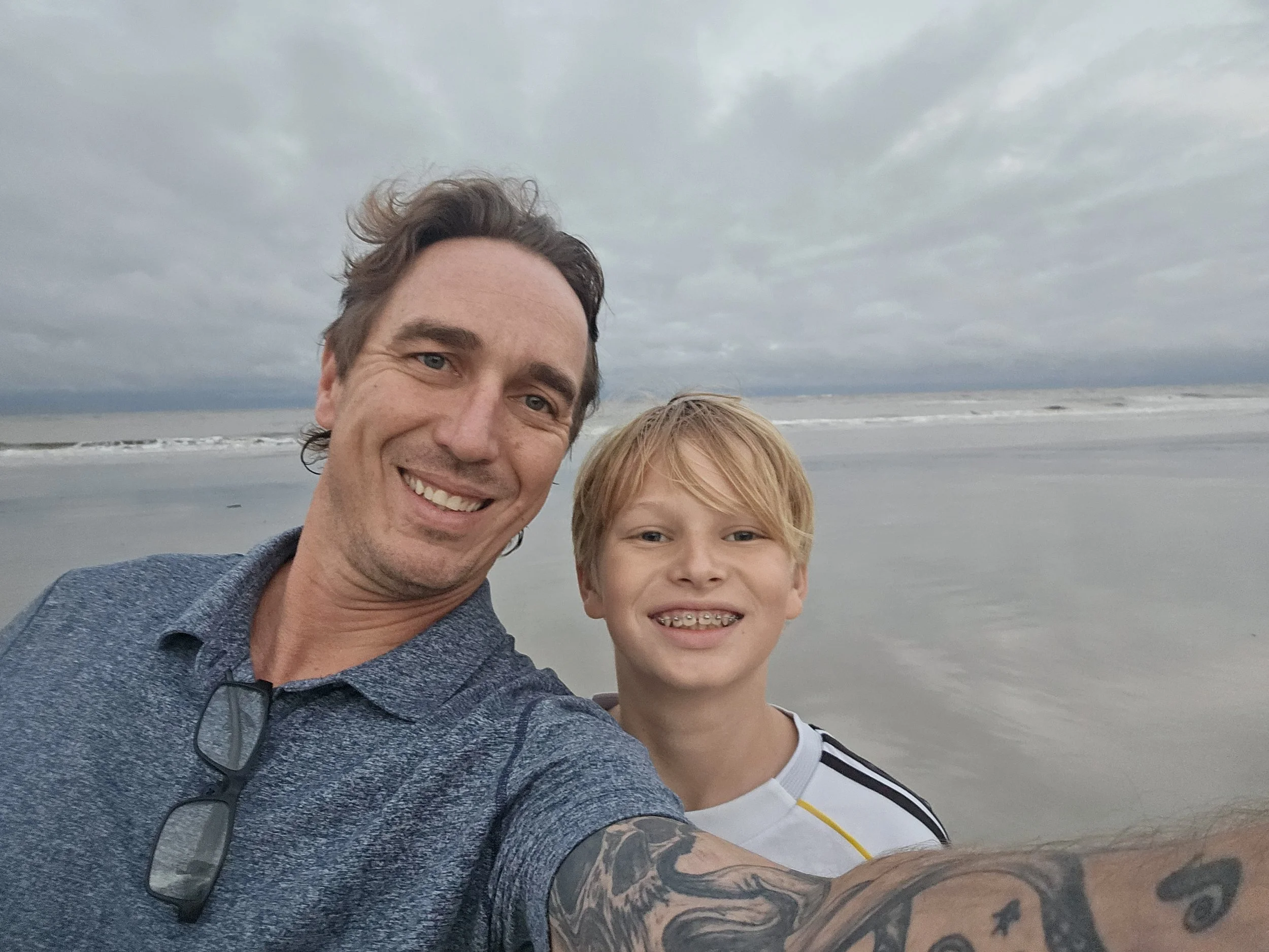 A man and a boy taking a selfie at the beach during cloudy weather, with the ocean and waves in the background.