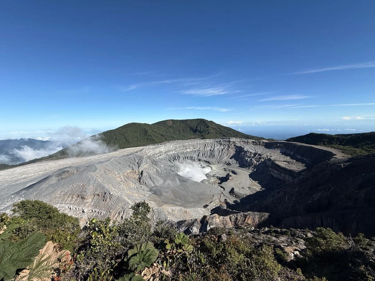 Volcanic crater with smoke and ash, surrounded by green hills under a blue sky.