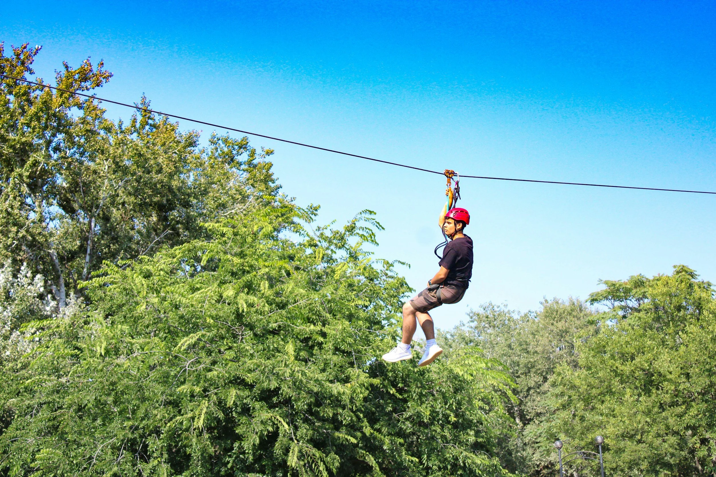 A person wearing a red helmet and black shirt riding a zip line over green trees under a clear blue sky.