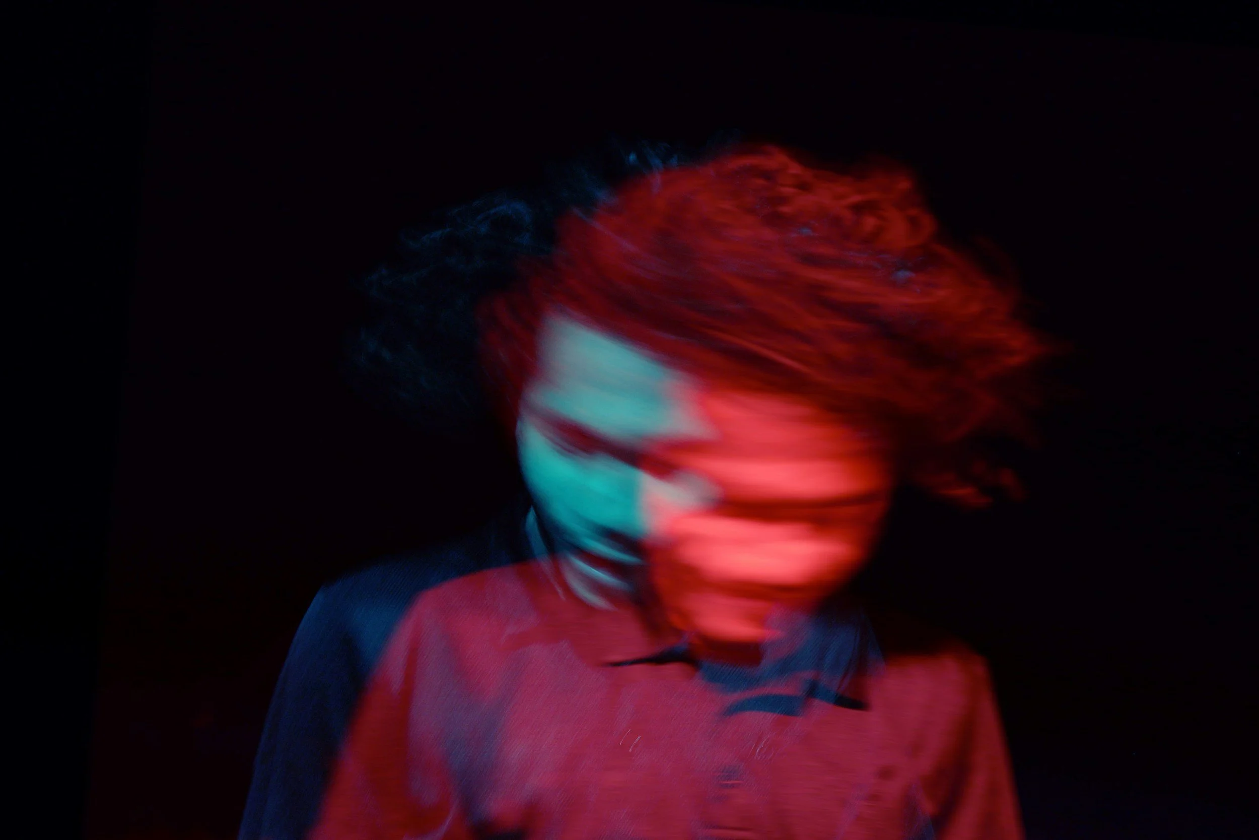 Blurred photo of a person with red hair moving swiftly, wearing a dark and red outfit, against a black background.