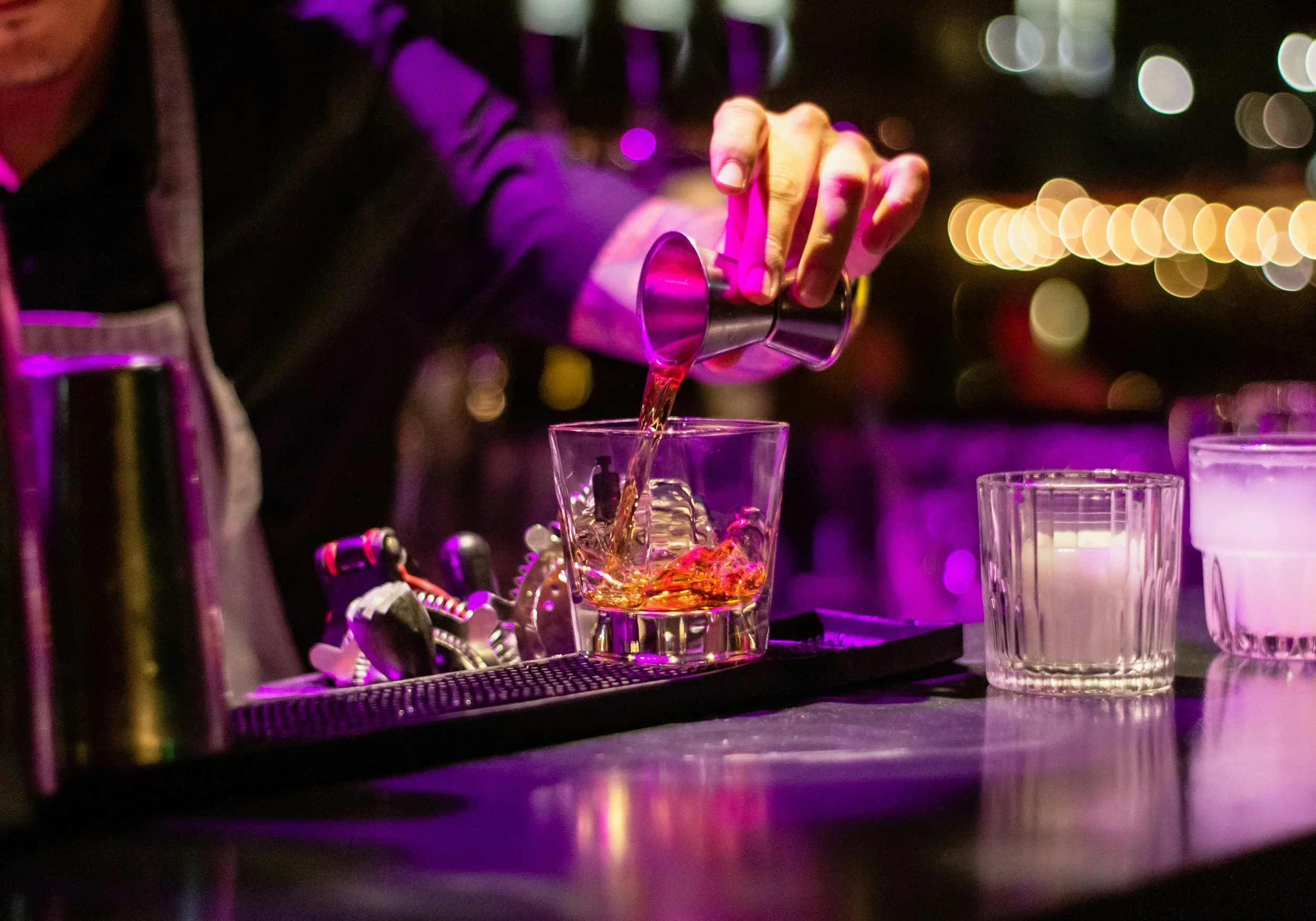 A bartender pouring a drink into a glass at a bar with colorful lighting and blurred bokeh lights in the background.