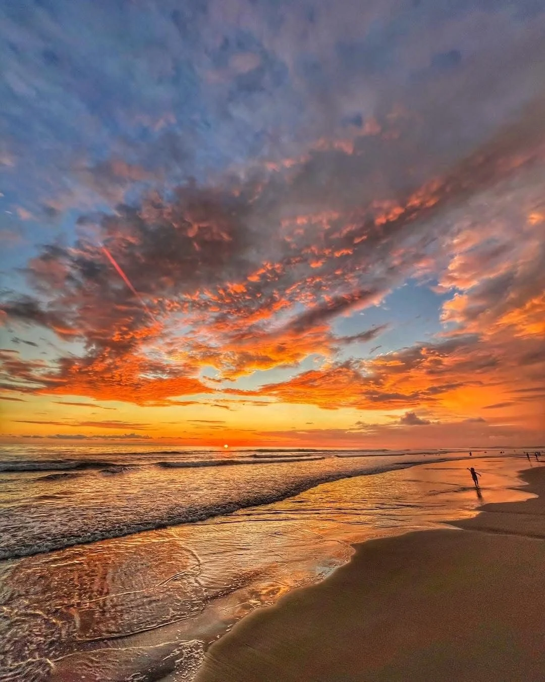 Sunset over a beach with colorful clouds and a few people walking near the water.