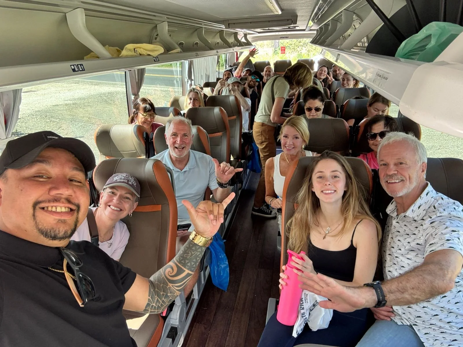 Inside a bus filled with smiling passengers and crew, with some people taking selfies and others interacting, suggesting they are traveling together on a group trip.