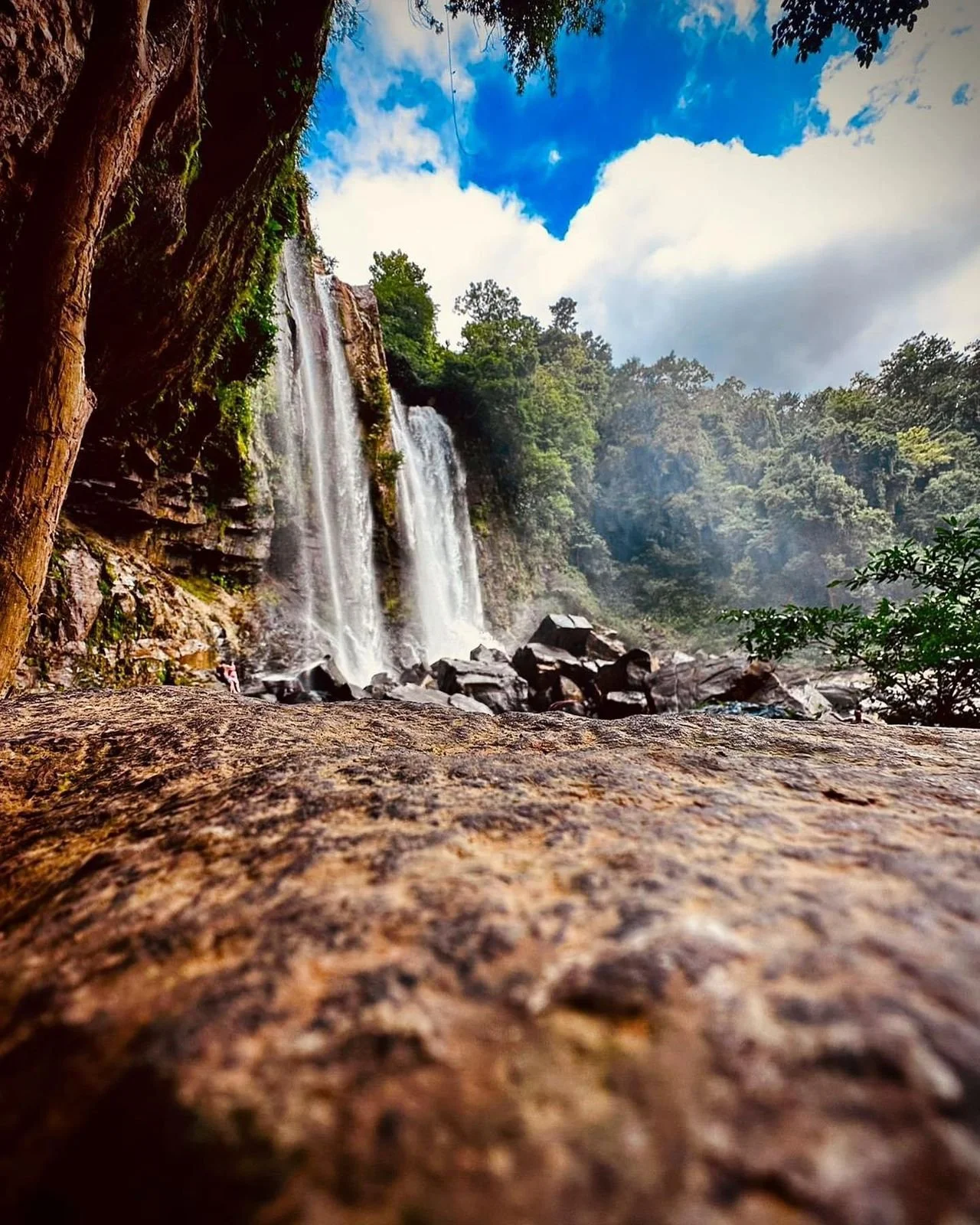 A scenic view of a waterfall cascading down a rocky cliff, surrounded by lush green trees under a partly cloudy blue sky.