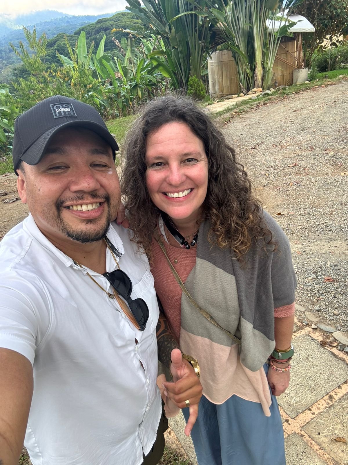 Smiling man and woman taking a selfie outdoors with lush green plants and mountains in the background.