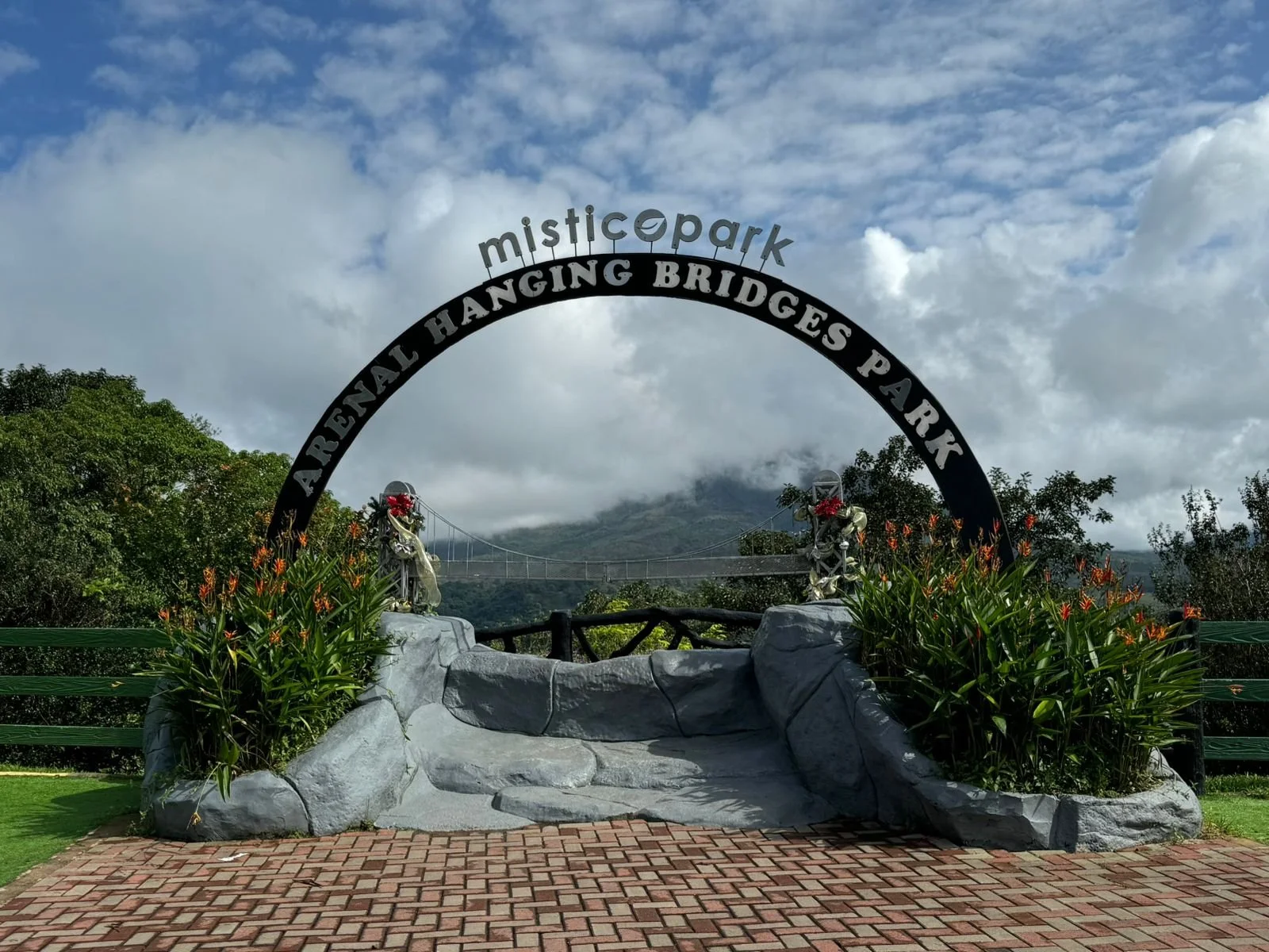 Entrance sign for Mistic Park with a black arch that reads 'Arretneal Hanging Bridges Park' and a metal arch saying 'mistic park', with a mountain and clouds in the background. There are decorative flowers and rocks at the base of the sign.