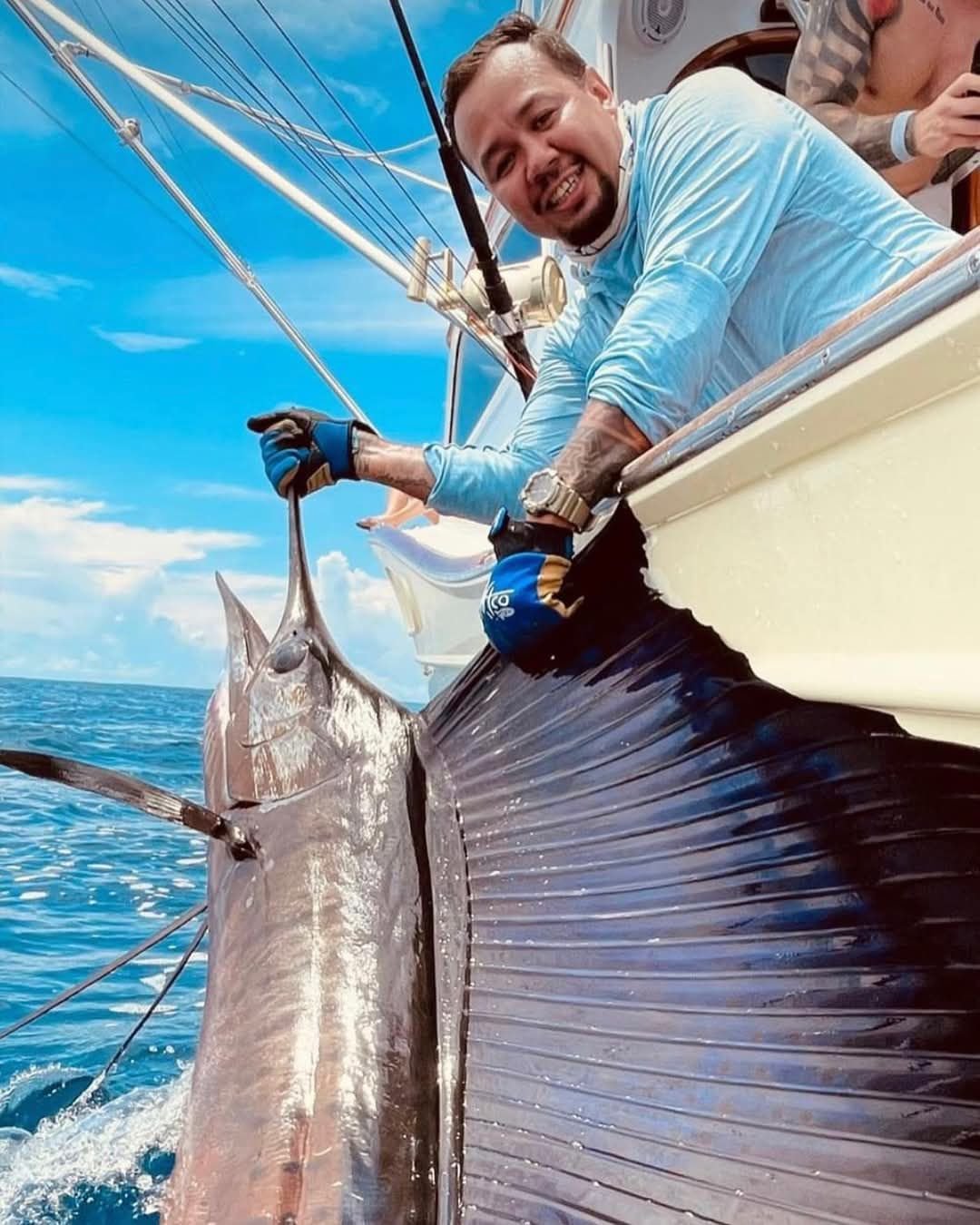 A man fishing from a boat, holding a large fish with one hand, smiling at the camera against a backdrop of blue sky and ocean.