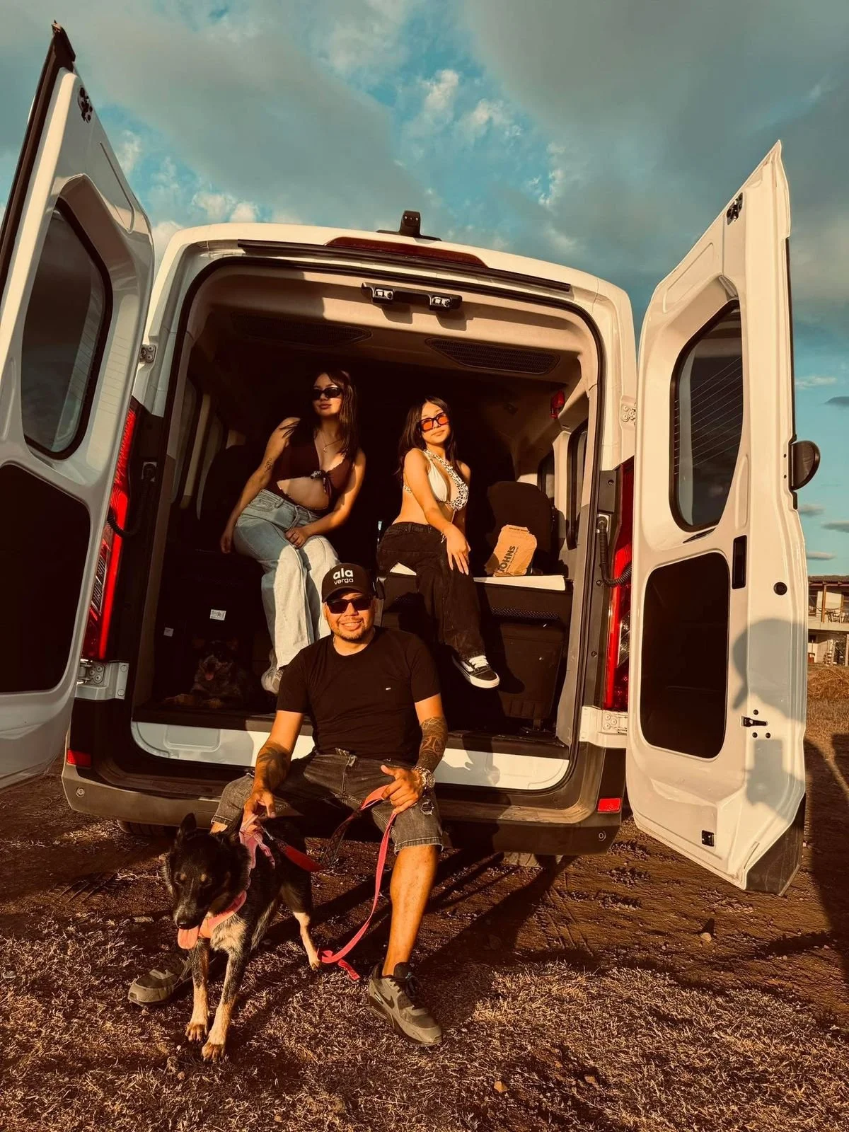 A group of four people and a dog are posing near a white van with open doors. Two women are sitting inside the van, and a man is sitting on the ground outside, holding a black dog on a pink leash. The scene is outdoors during sunset with a partly cloudy sky.