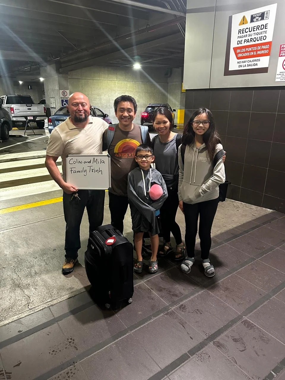 Group of five people standing together in an outdoor parking garage, with a man holding a whiteboard that says 'Colin and Mila Family Trip.' There is a suitcase in front of them. The group appears to be happy, possibly leaving for or returning from a