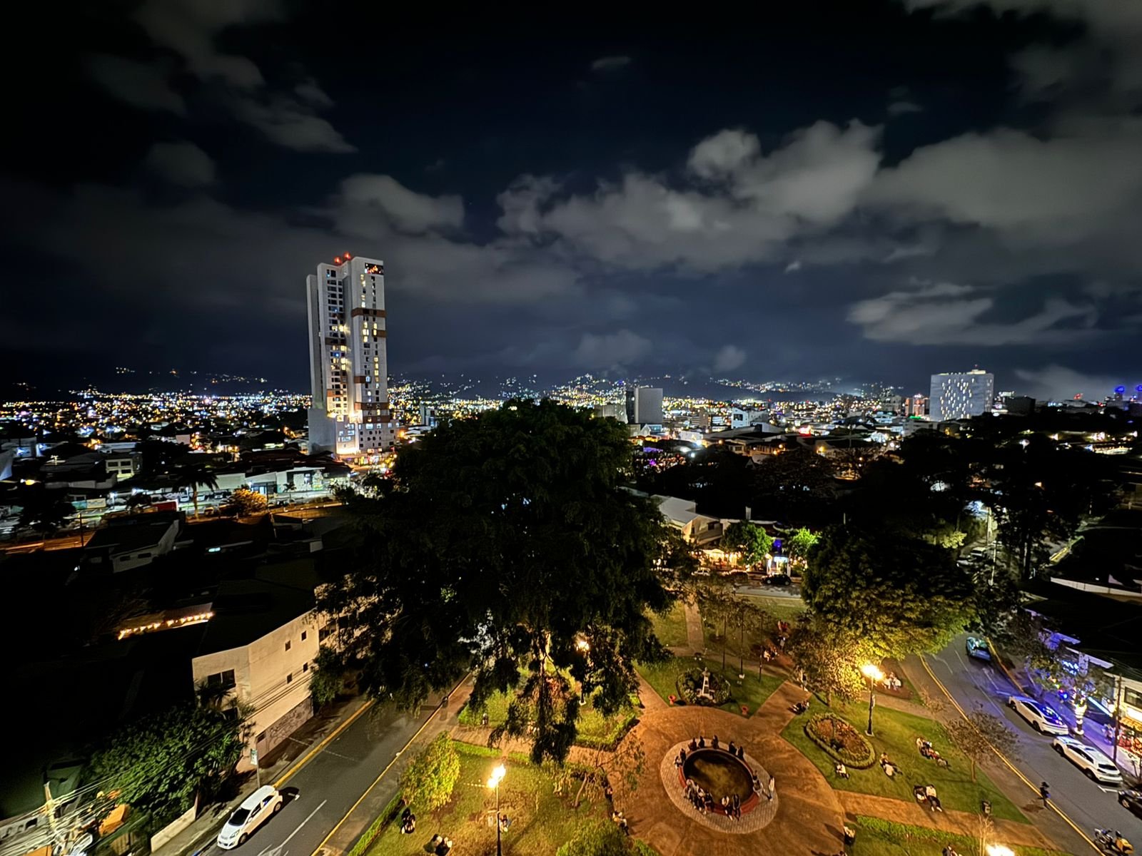 Nighttime cityscape with a tall illuminated building, streets lined with trees, parked cars, and a park with a fountain in the foreground. Clouds are in the sky, and city lights extend to the horizon.