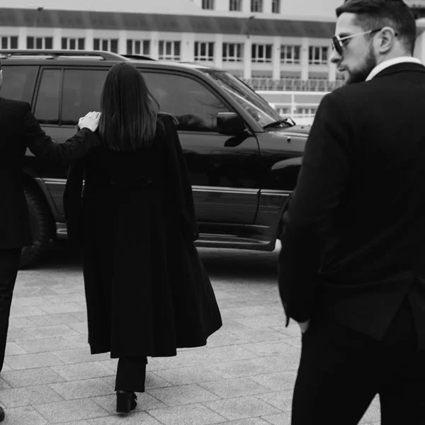 Three people in formal attire standing near a black SUV outdoors, with a city building in the background.