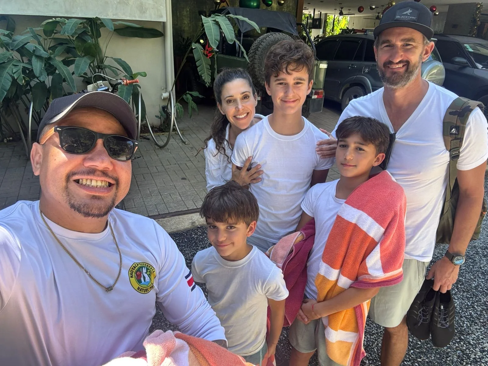 Group of seven people, including three children and four adults, smiling for a selfie outside a building with cars and bicycles in the background.