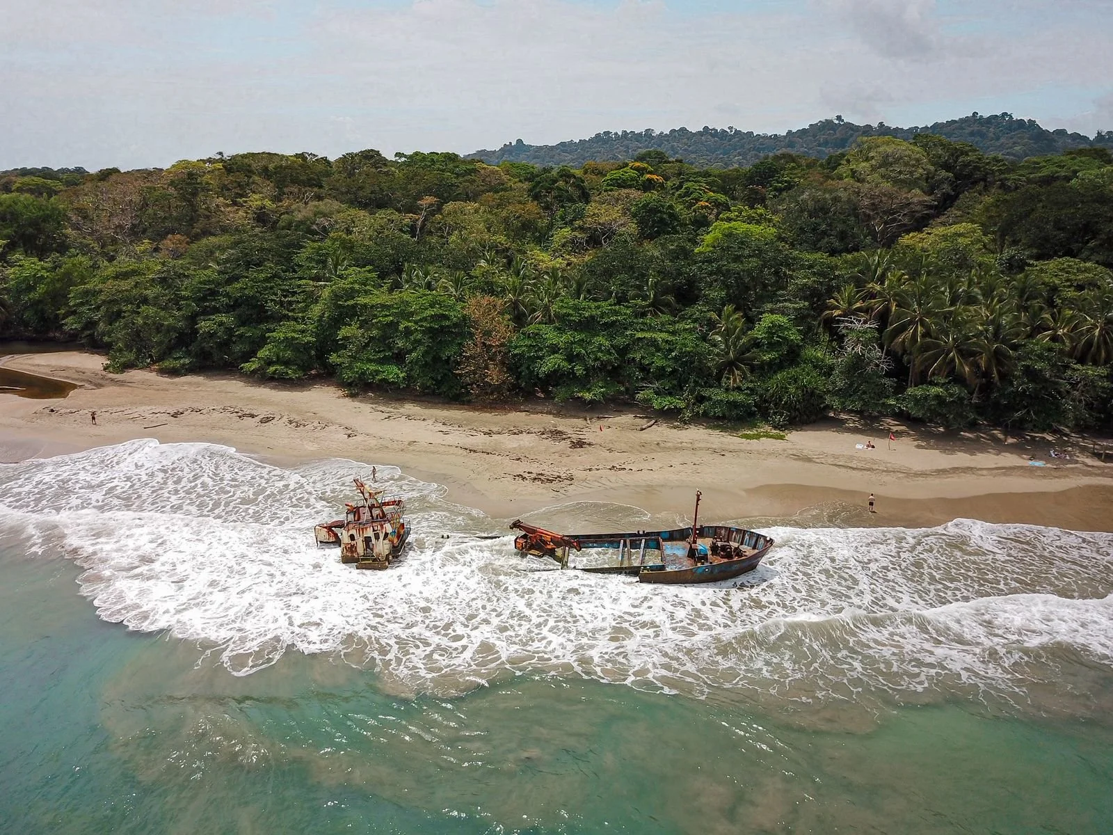 An abandoned shipwreck washed ashore on a sandy beach with lush green trees and hills in the background.