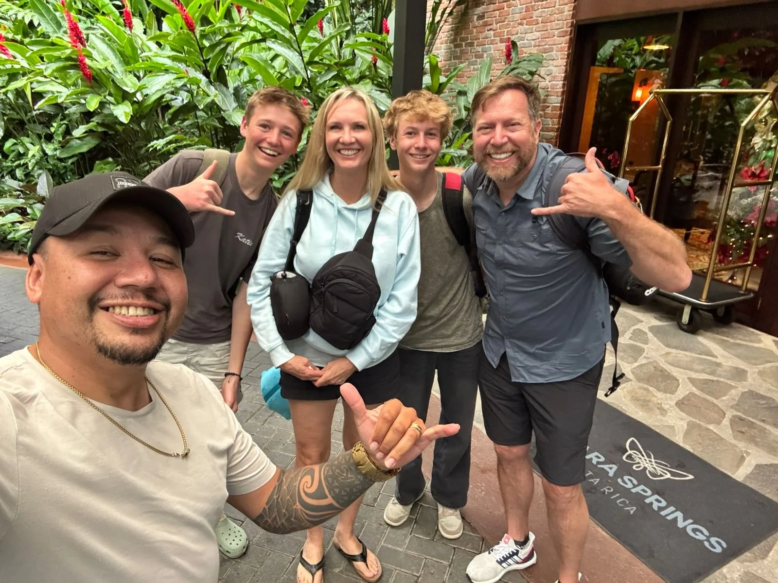 A group of six people smiling and posing for a photo outside a hotel entrance with lush greenery and a floral display in the background. The person in front is taking a selfie, and the others are making gesture signs with their hands.