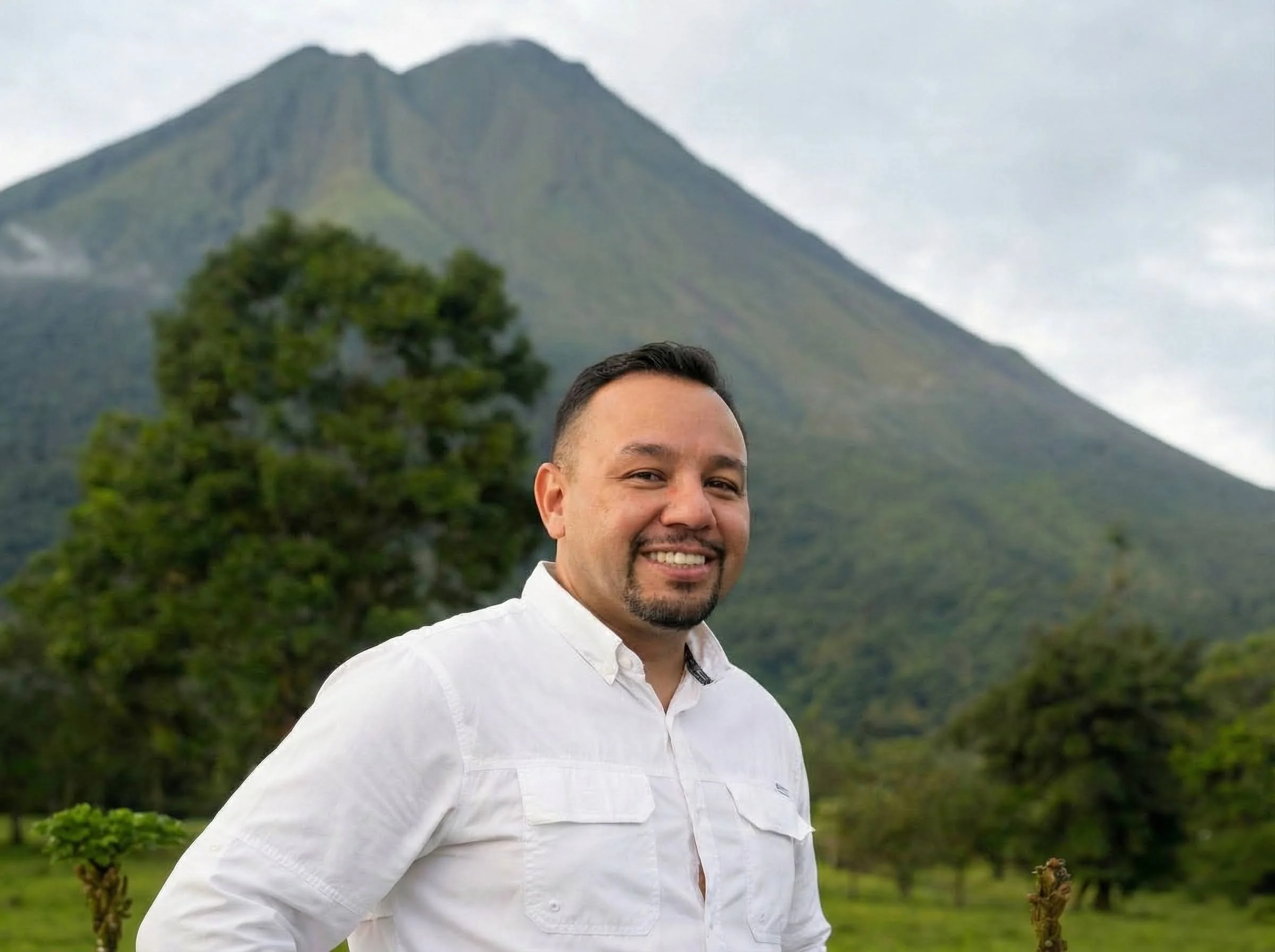 Smiling man in white shirt standing outdoors with lush green trees and mountains in the background.