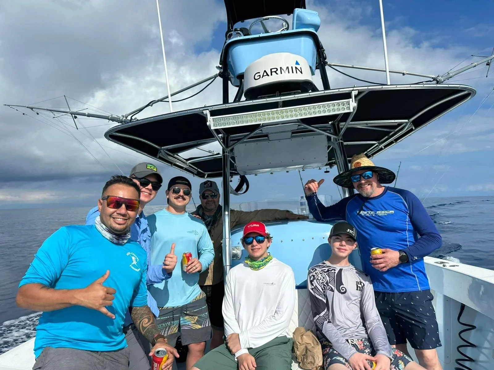 A group of eight people on a boat, wearing casual and outdoor clothing, some with sunglasses and hats, smiling and holding cans of drinks, with a blue ocean and cloudy sky in the background.