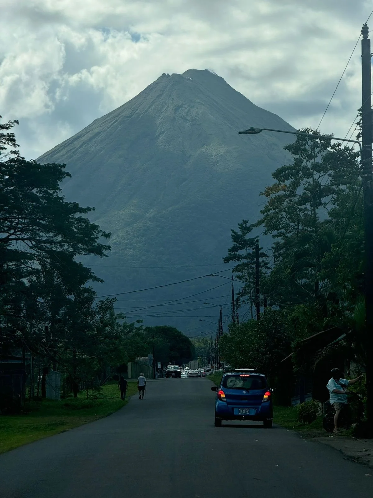 Street with trees and cars leading toward a volcano in the distance, under a cloudy sky.