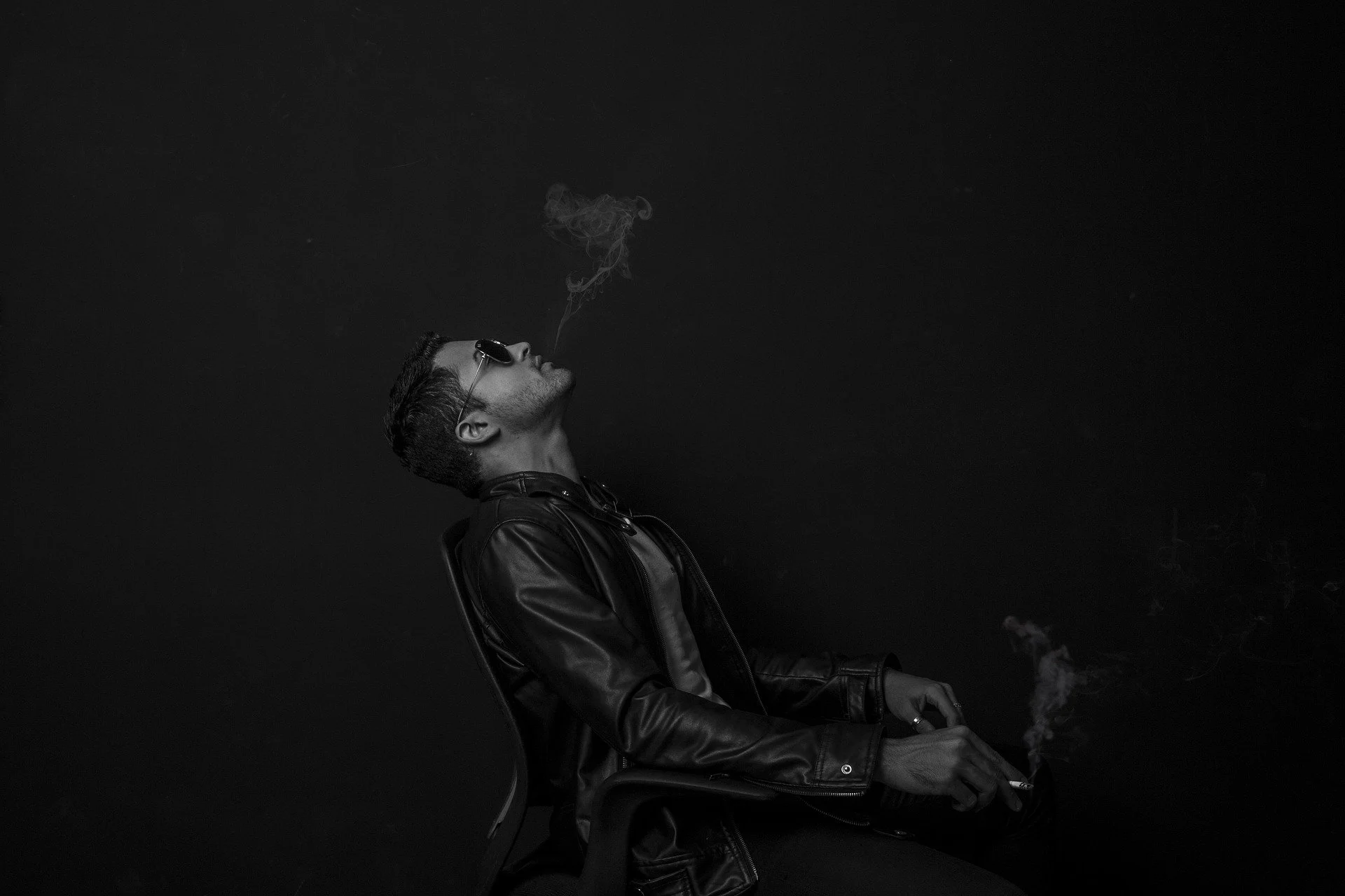A black and white photo of a young man with sunglasses in a leather jacket, sitting against a black background, exhaling smoke from a cigarette.