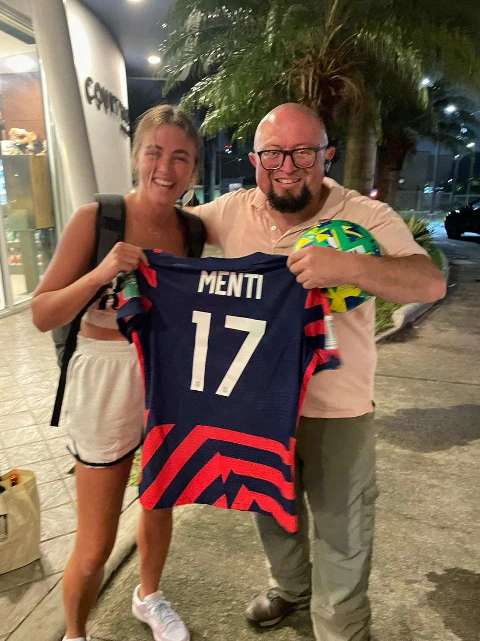A young woman and an older man smiling and holding a sports jersey with the name 'MENTI' and the number 17. The man is also holding a colorful soccer ball. They are standing outside at night near palm trees.