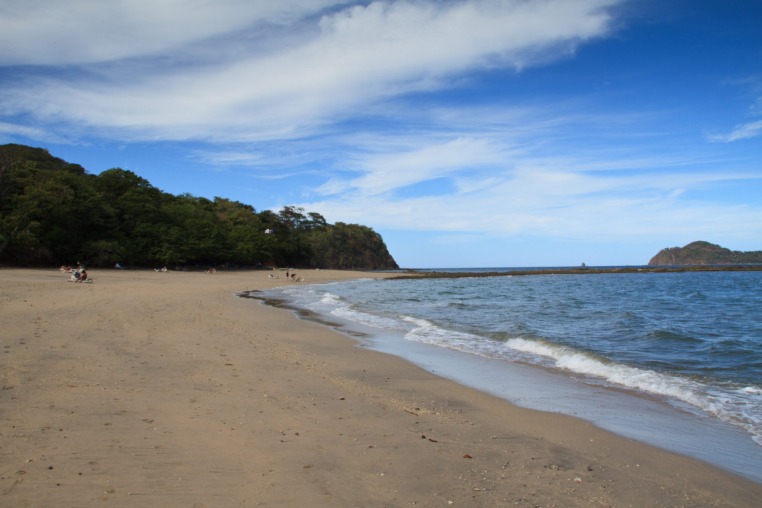 A peaceful beach with sand, blue water, and a partly cloudy sky. There are a few people and dogs relaxing on the beach near green, wooded hills in the background.