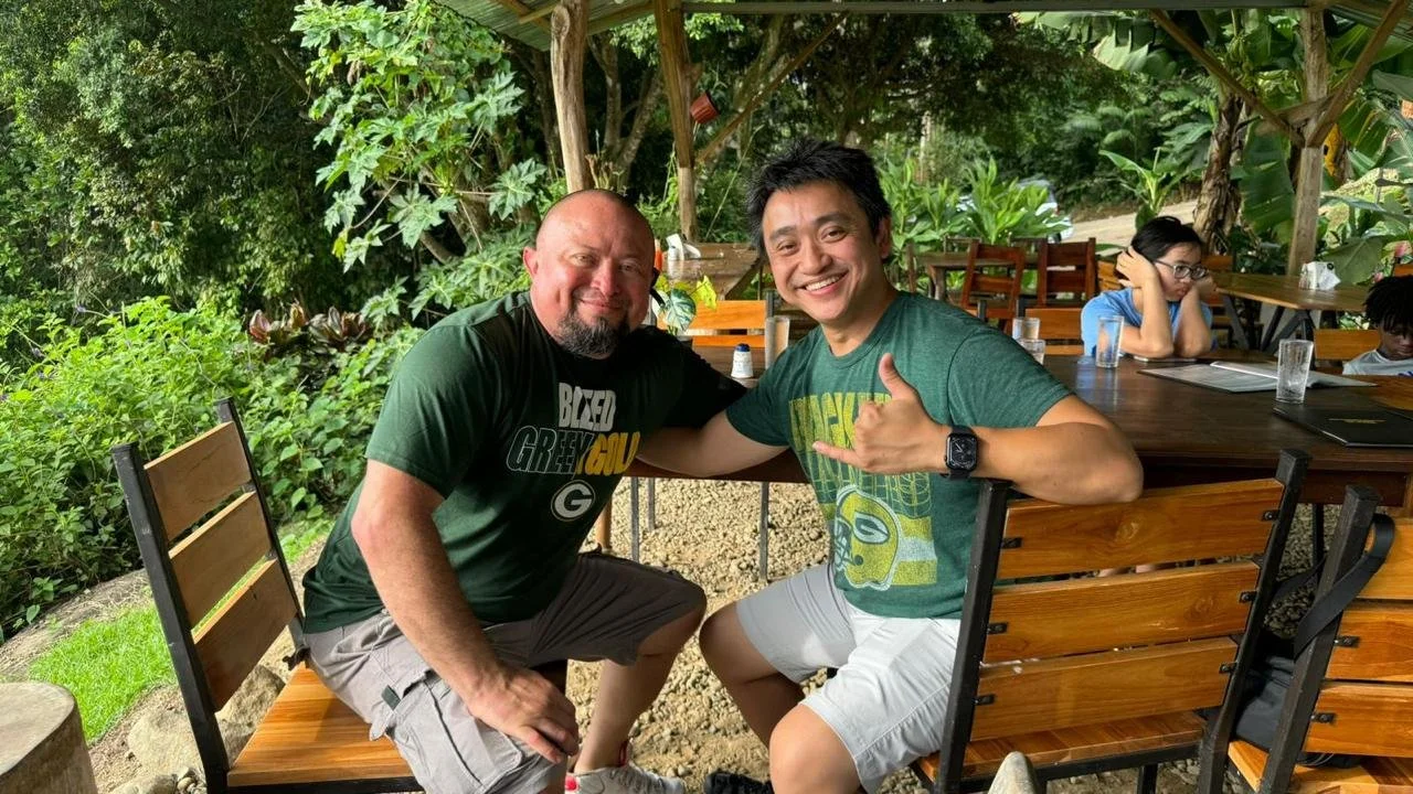 Two men smiling and sitting on wooden chairs outside, one making a shaka sign. Background with trees and other people at a seating area.