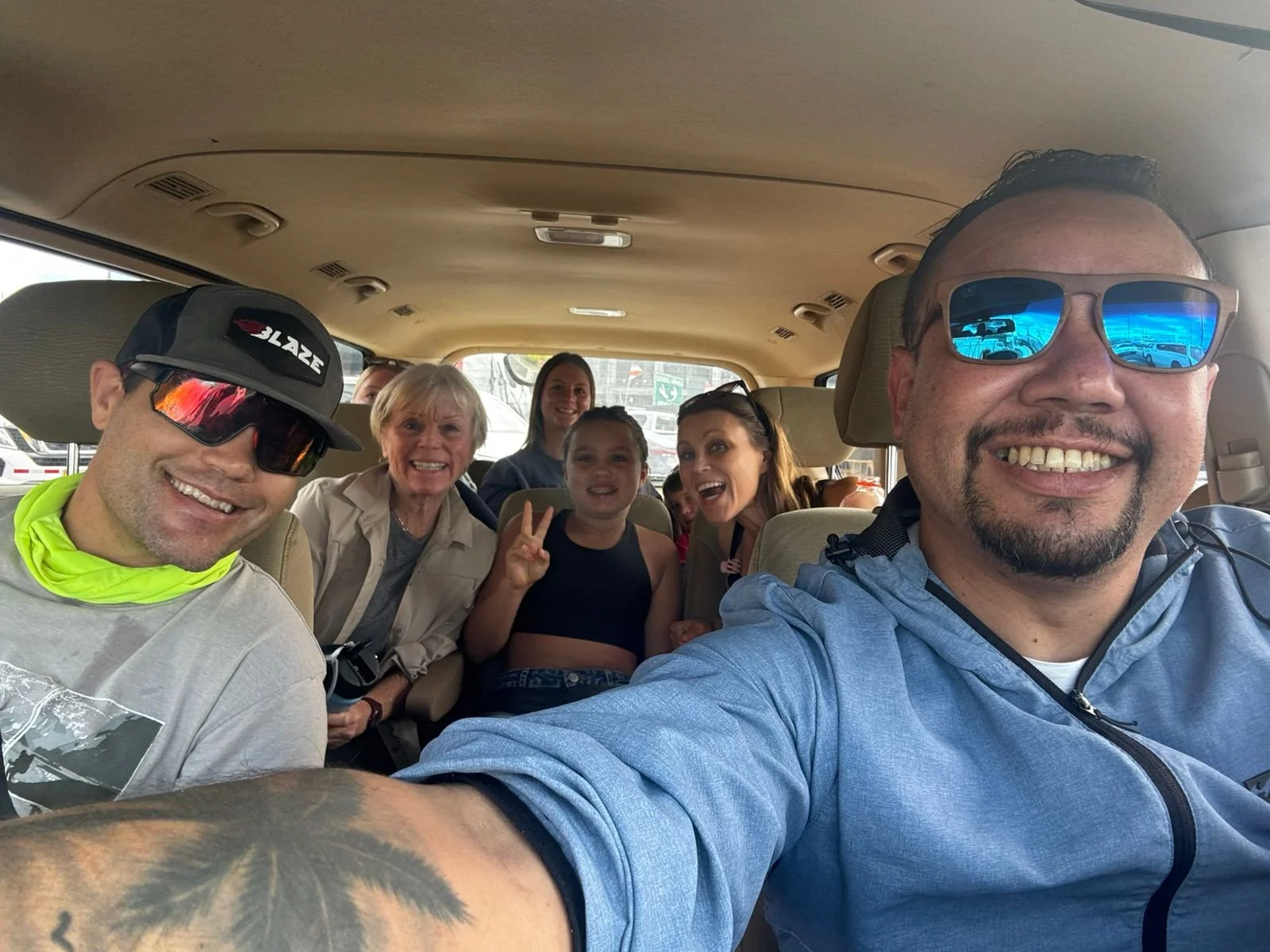 Group of seven people smiling and making peace signs inside a vehicle, with two adults wearing sunglasses in the front and five children in the back.