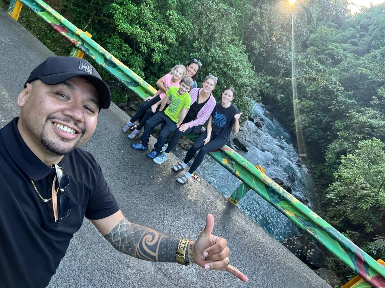 A group of six people sitting on a colorful green railing near a river, taking a selfie during the daytime with sunlight filtering through trees in the background.