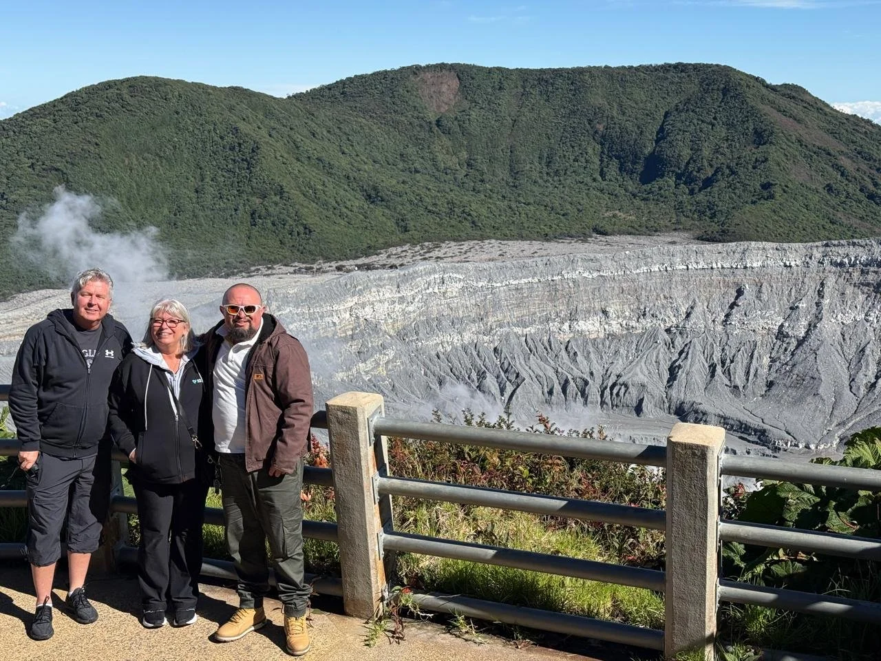Three people standing in front of a volcanic crater with green hills in the background, wearing casual outdoor clothing.