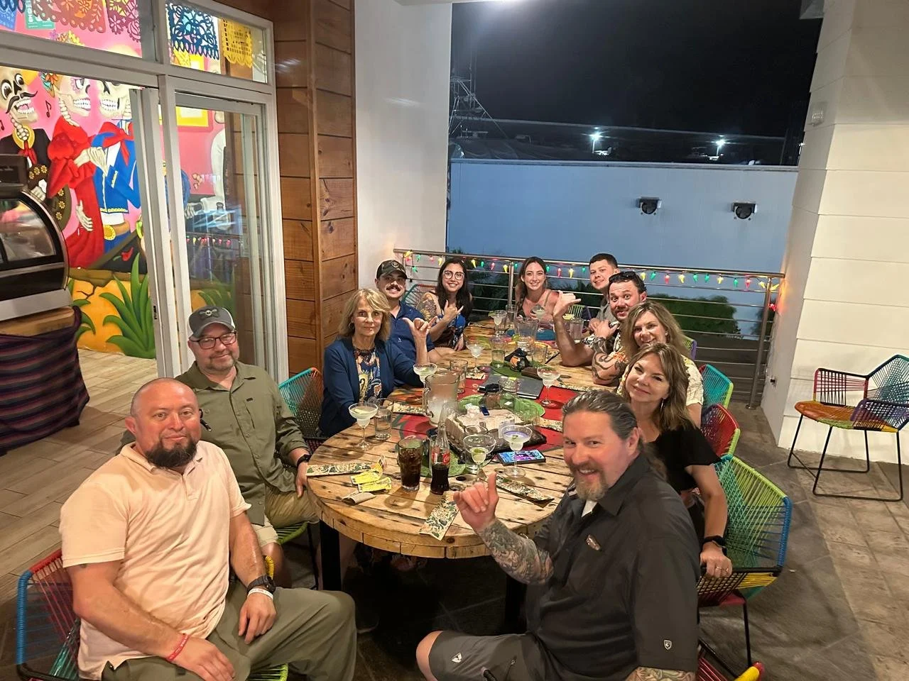 A group of twelve adults sitting around a wooden table at an outdoor restaurant or patio at night.