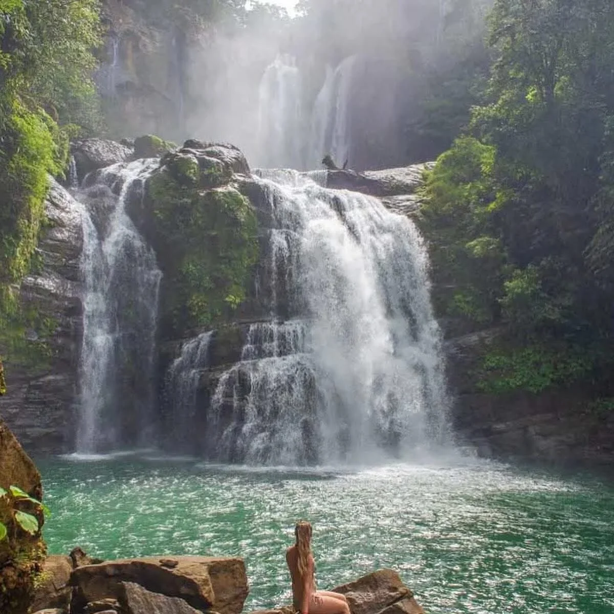 A scenic waterfall cascading over rocks and surrounded by lush greenery, with a woman sitting on a rock in the foreground.