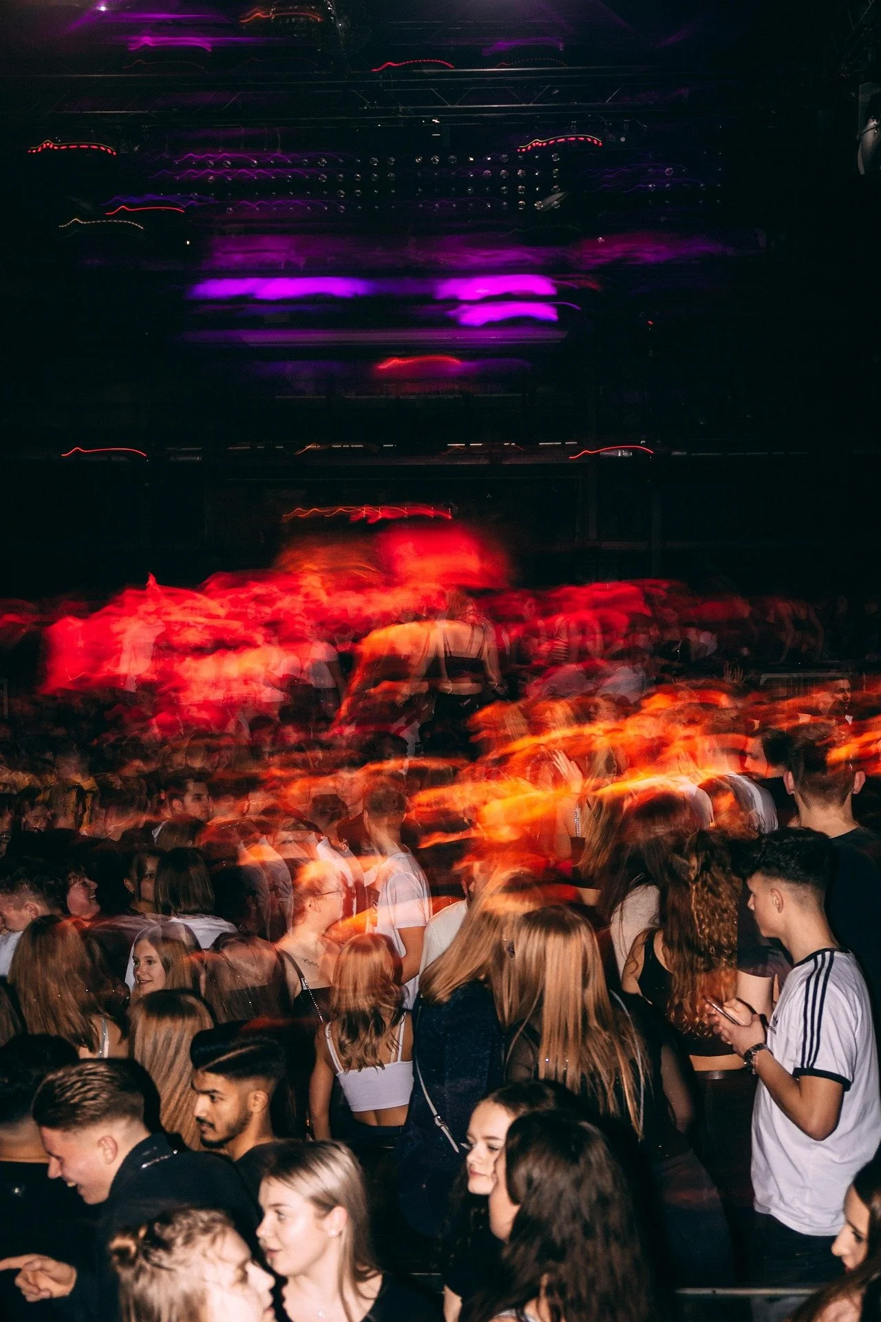 Crowd of people at a concert or nightclub, with colorful red and purple lighting effects and motion blur.