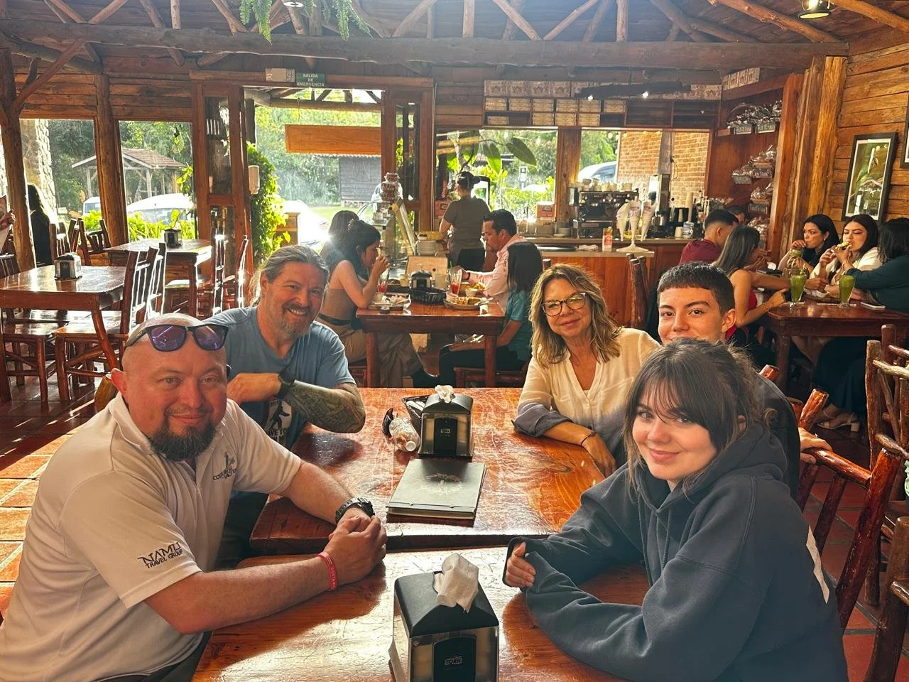 Group of five people sitting at a wooden table in a cozy restaurant or cafe, smiling for the camera, with other patrons and a coffee station in the background.