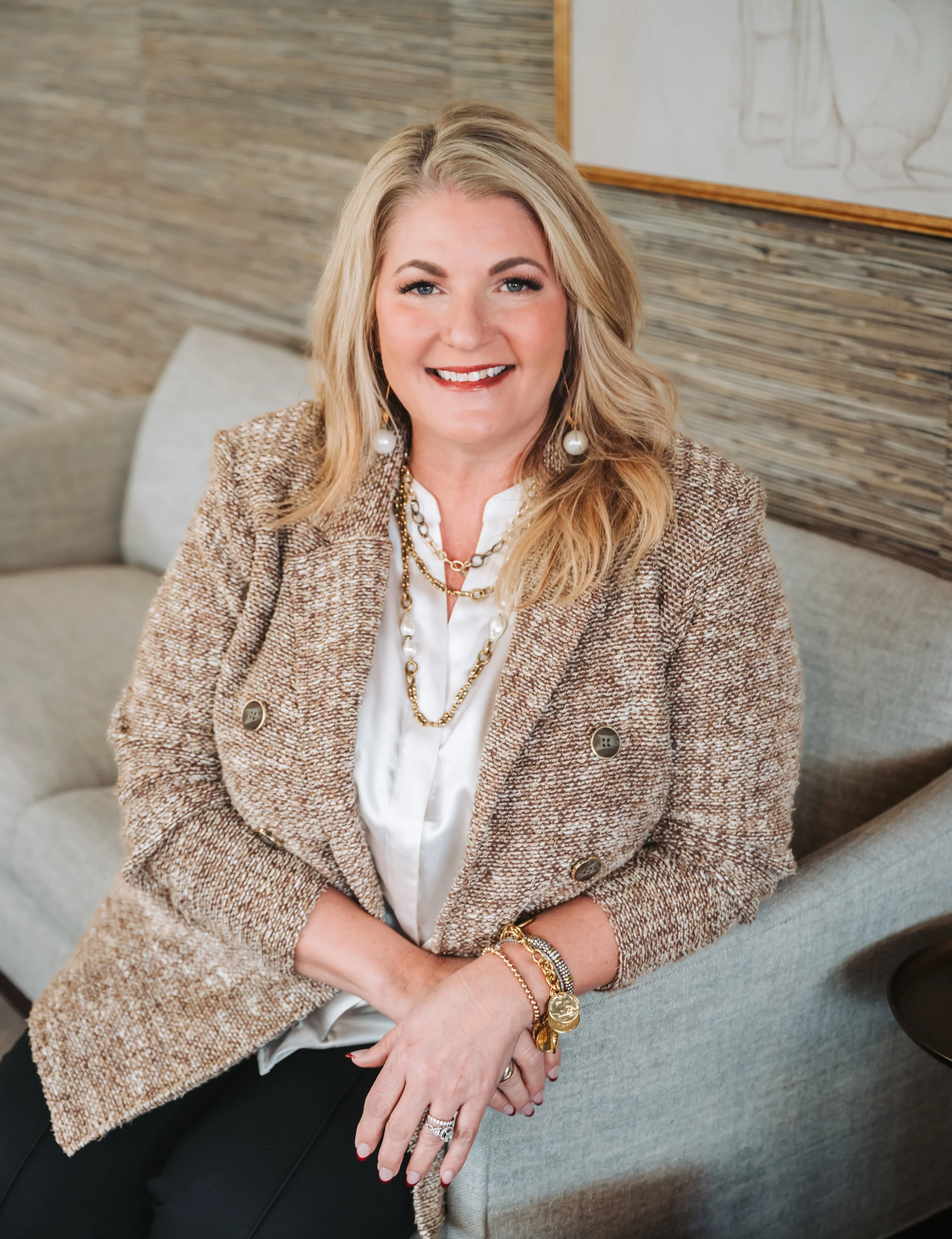 A woman with blonde hair, smiling, wearing a gold jewelry set including earrings, necklaces, bracelets, and rings, dressed in a tweed blazer and white shirt, sitting on a gray couch in a modern interior with a wood and stone accent wall.