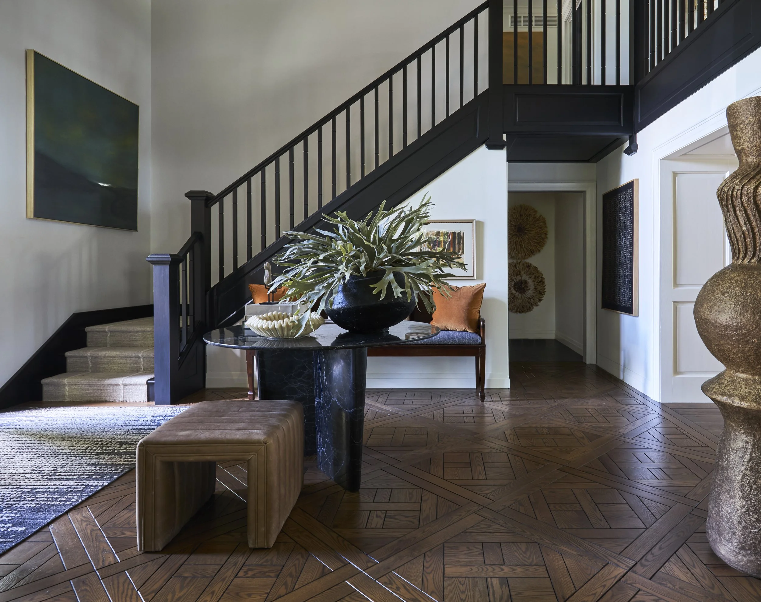 Tudor style foyer with custom parquet flooring and wood stair railing. Dark stained wood newel post and balusters against light walls. Foyer table with green foliage in a vase. Bench with pillow against staircase wall. Large artwork on stair landing.