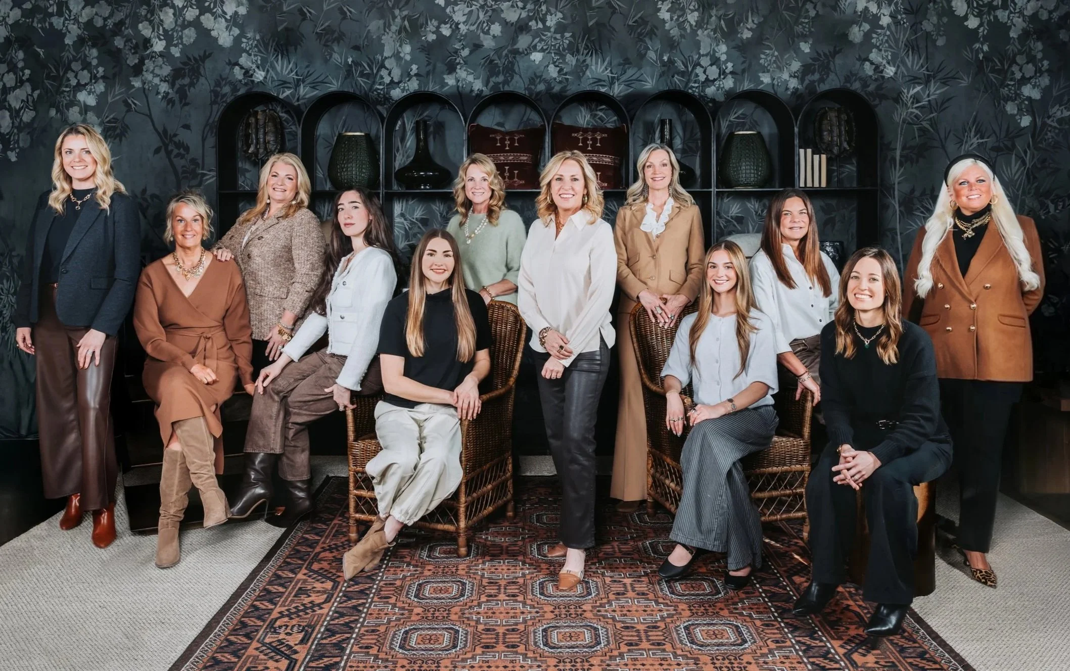 A group of fifteen women posing together in a living room, some seated and others standing, with a decorative dark wall and shelving behind them.