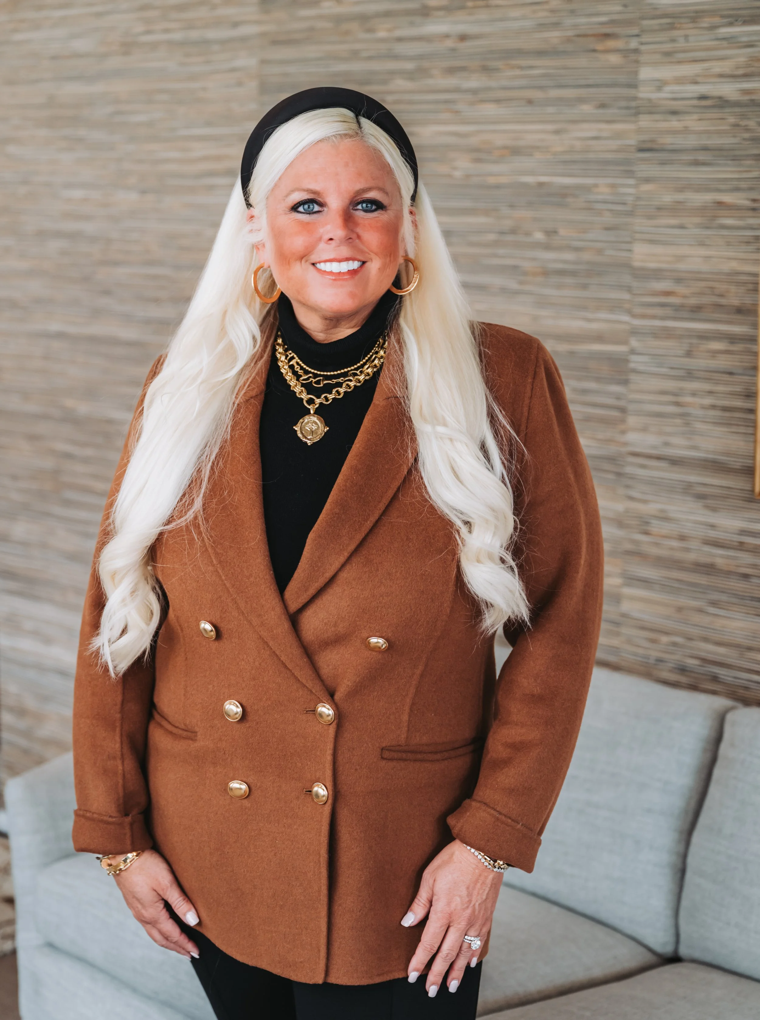 A woman with long white hair, wearing a black headband, gold jewelry, and a brown double-breasted coat, standing outdoors.