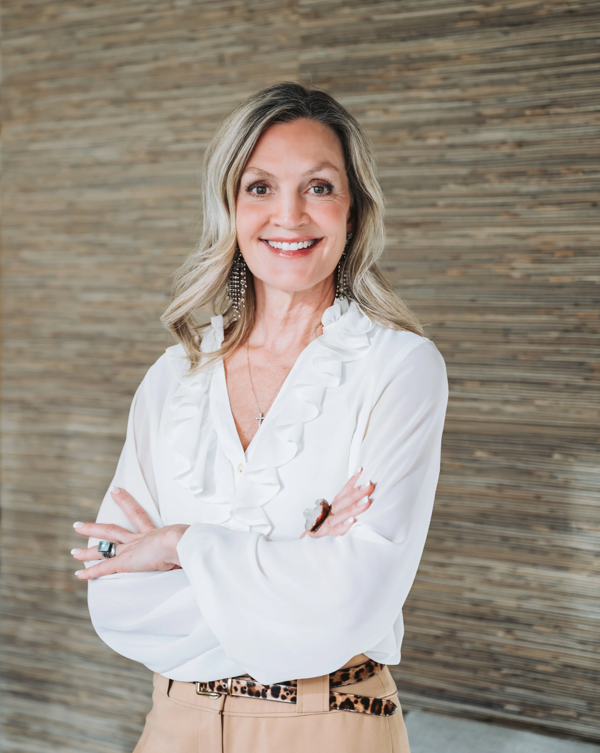 A woman with blonde hair smiling, wearing a white blouse, leopard-print belt, and large earrings, standing in front of a textured horizontal wooden wall.