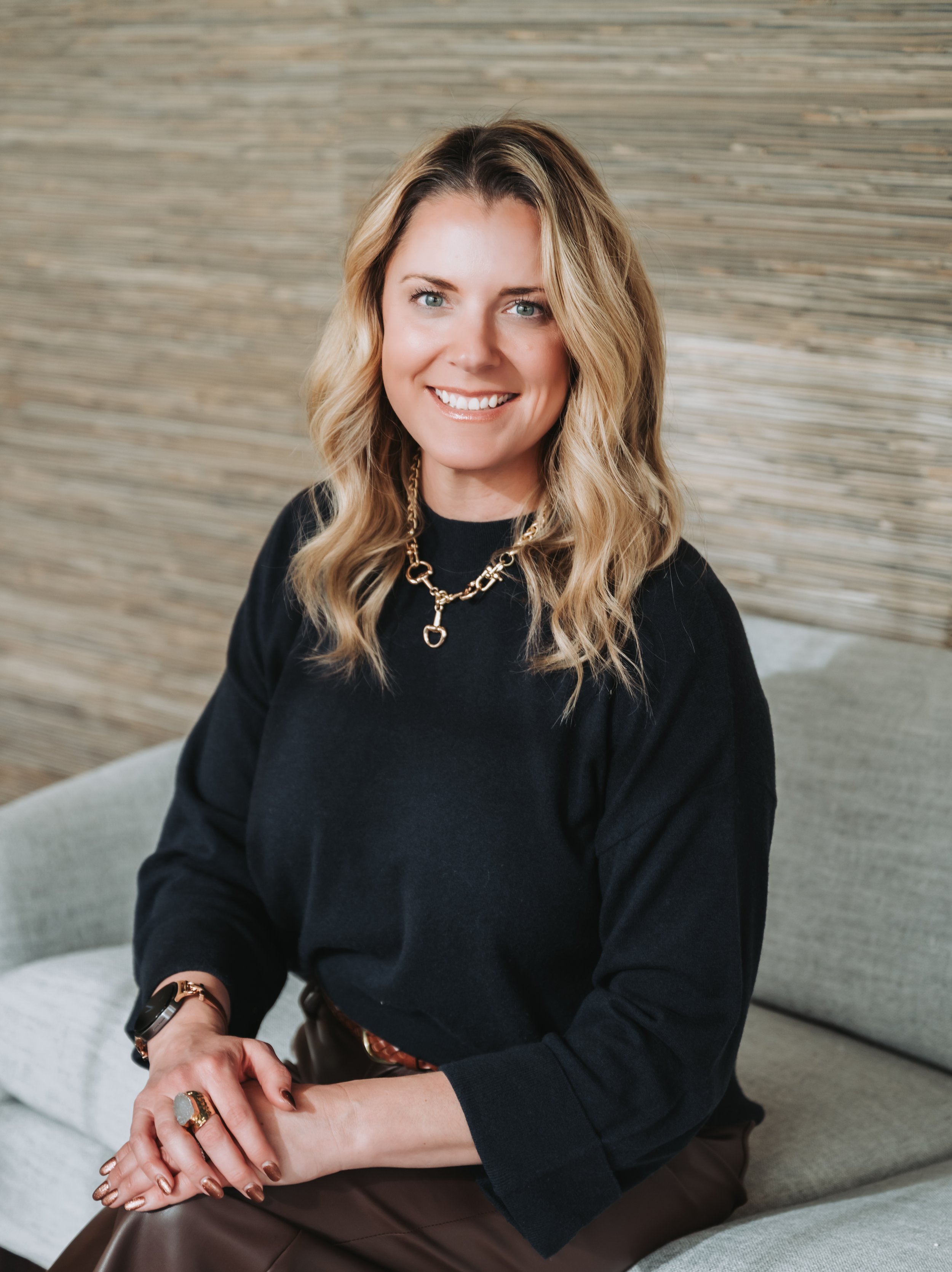 Smiling woman with blonde wavy hair sitting on a gray couch, wearing a black top and gold jewelry, with a textured wooden wall in the background.