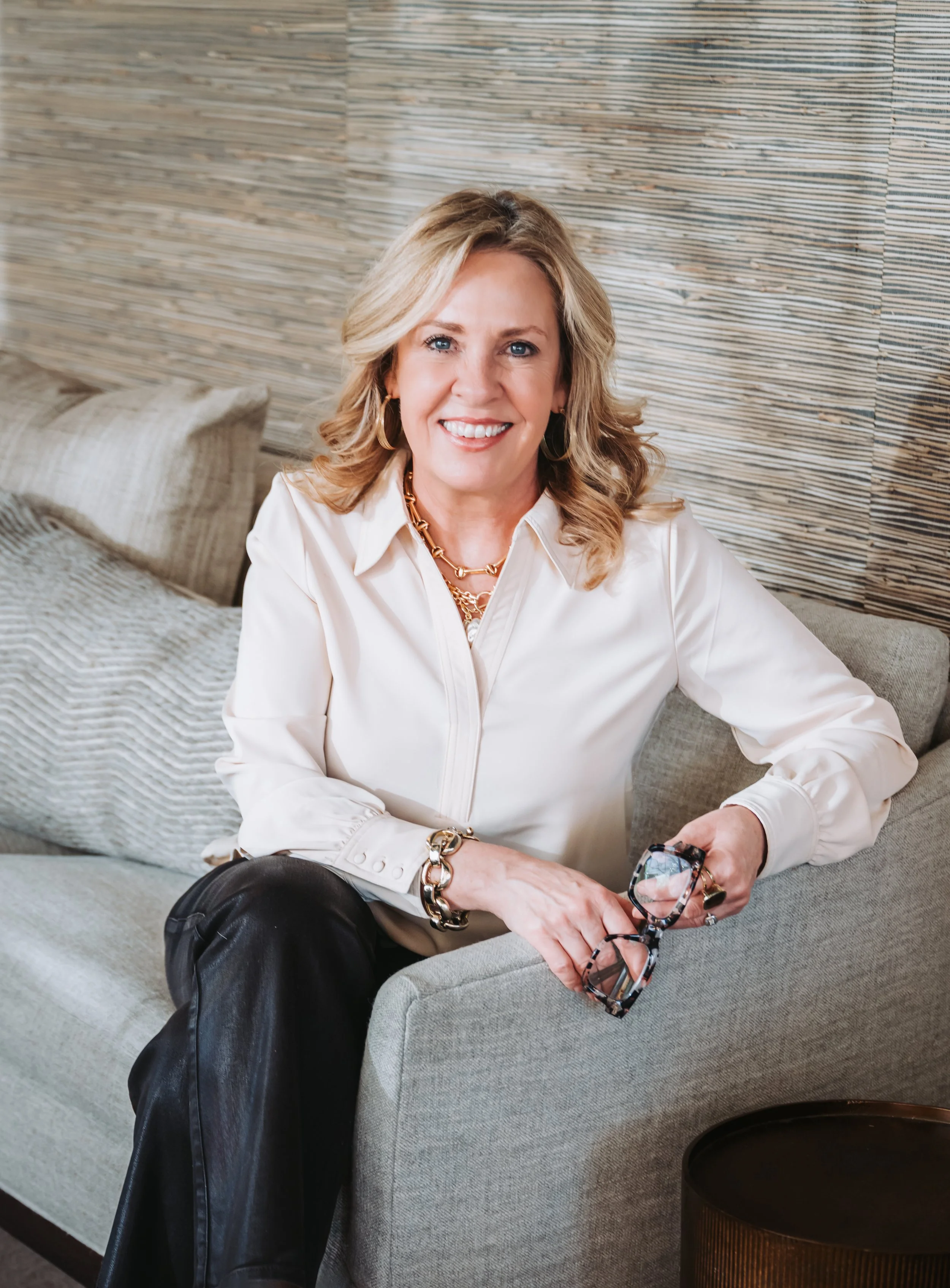 A woman with blonde hair, blue eyes, and wearing a white blouse, gold jewelry, and holding glasses, sitting on a light-colored sofa with a textured beige pillow and a woven wooden wall behind her.