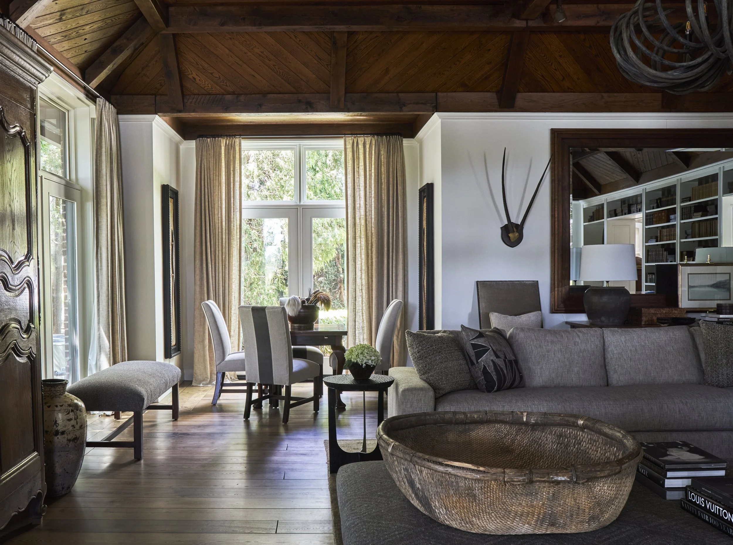 Great room with game table near windows. Beams on ceiling with tongue and groove. Tudor style architecture. Hardwood flooring and floor to ceiling windows. Antlers on wall.