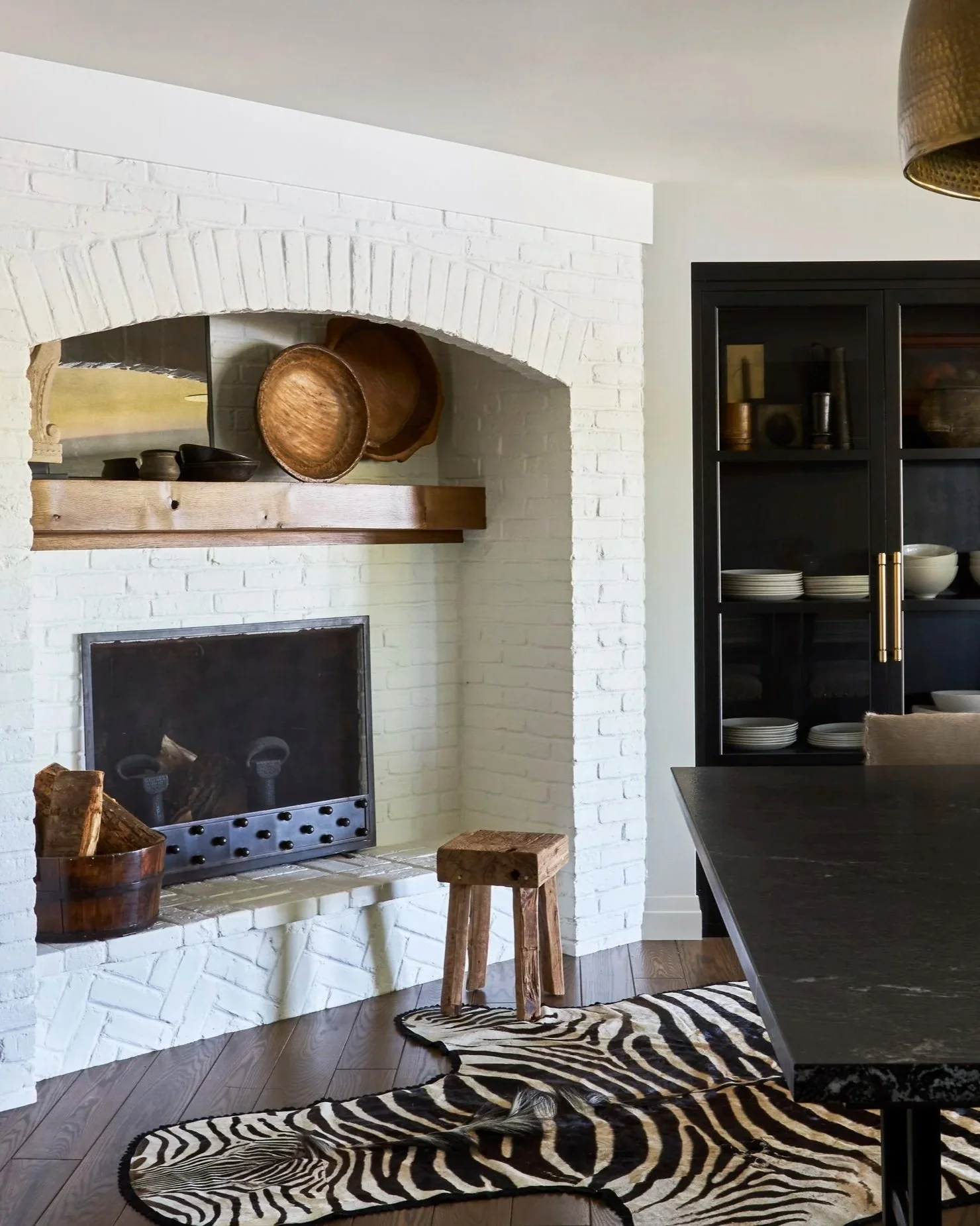 white brick fireplace in kitchen. Zebra hyde rug. Dish armoire inset into wall. small stool in front of fireplace.