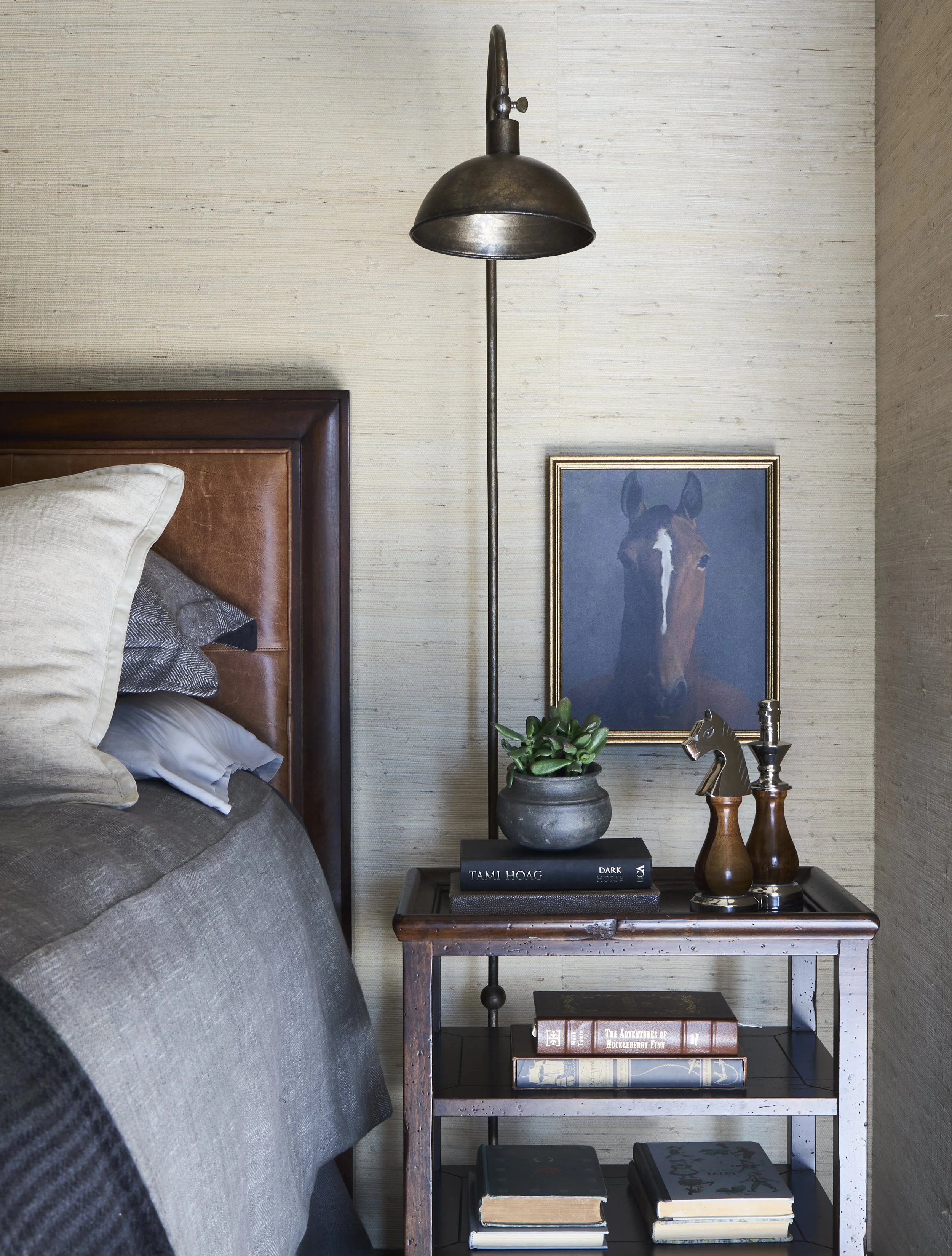 grasscloth in bedroom, leather headboard, horse decor, nightstand with books, artwork by bed. Sconce lighting next to bed.