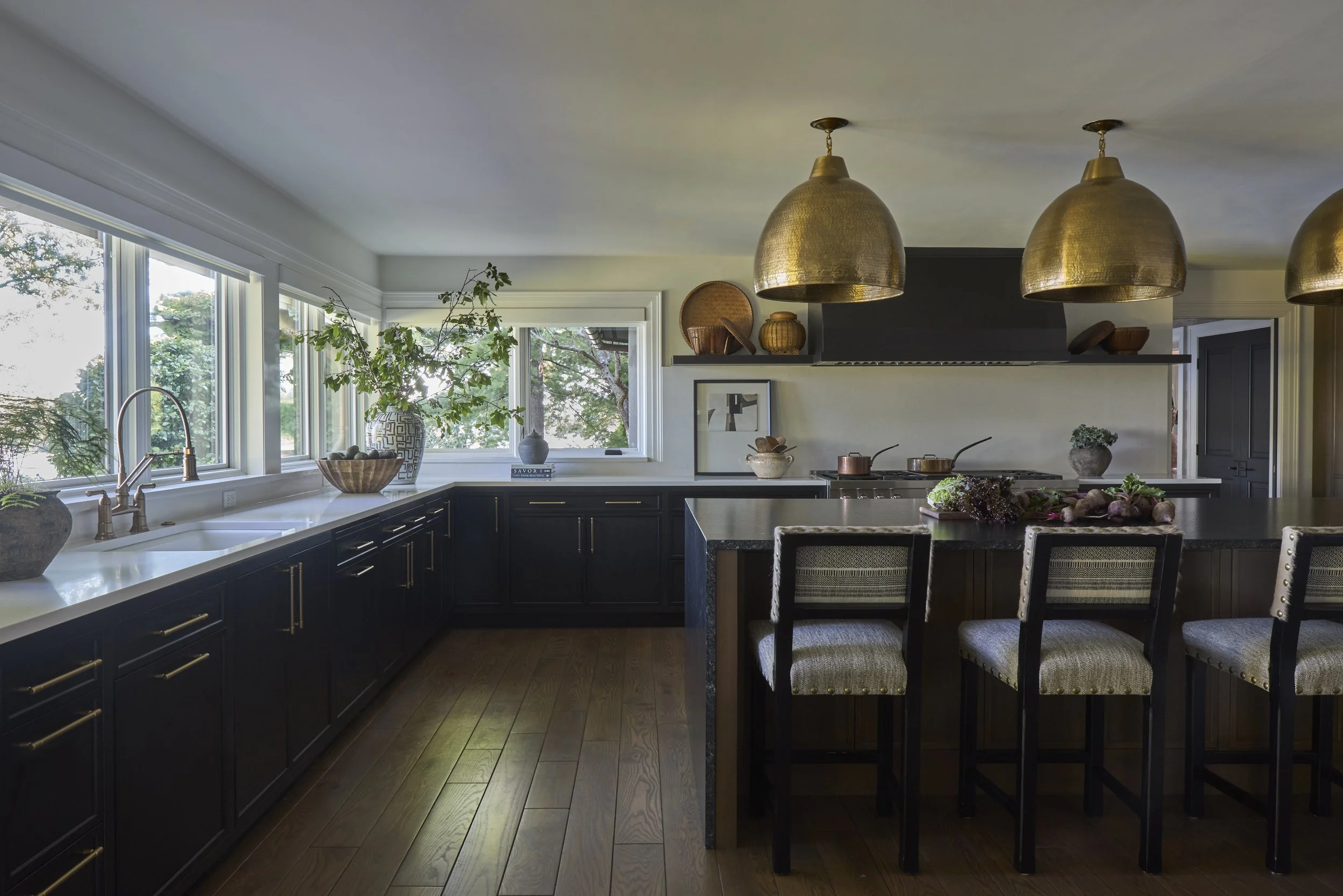 L-shaped kitchen with large windows, island with seating. Large gold pendants over island. Upholstered counter stools with nailheads. Island with waterfall sides. Dark cabinets with light countertops.