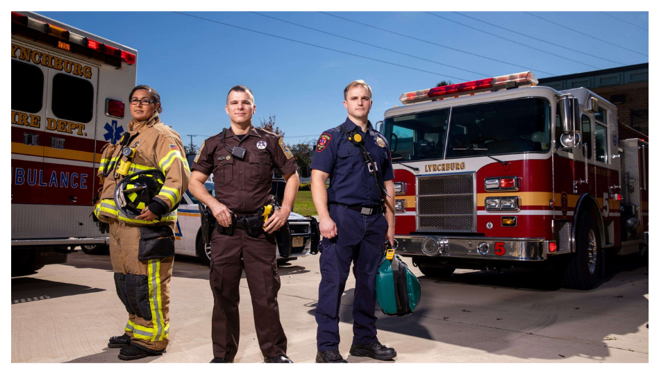 Three emergency responders, including a firefighter in turnout gear, a police officer, and a paramedic, stand in front of an ambulance and a fire truck on a sunny day.