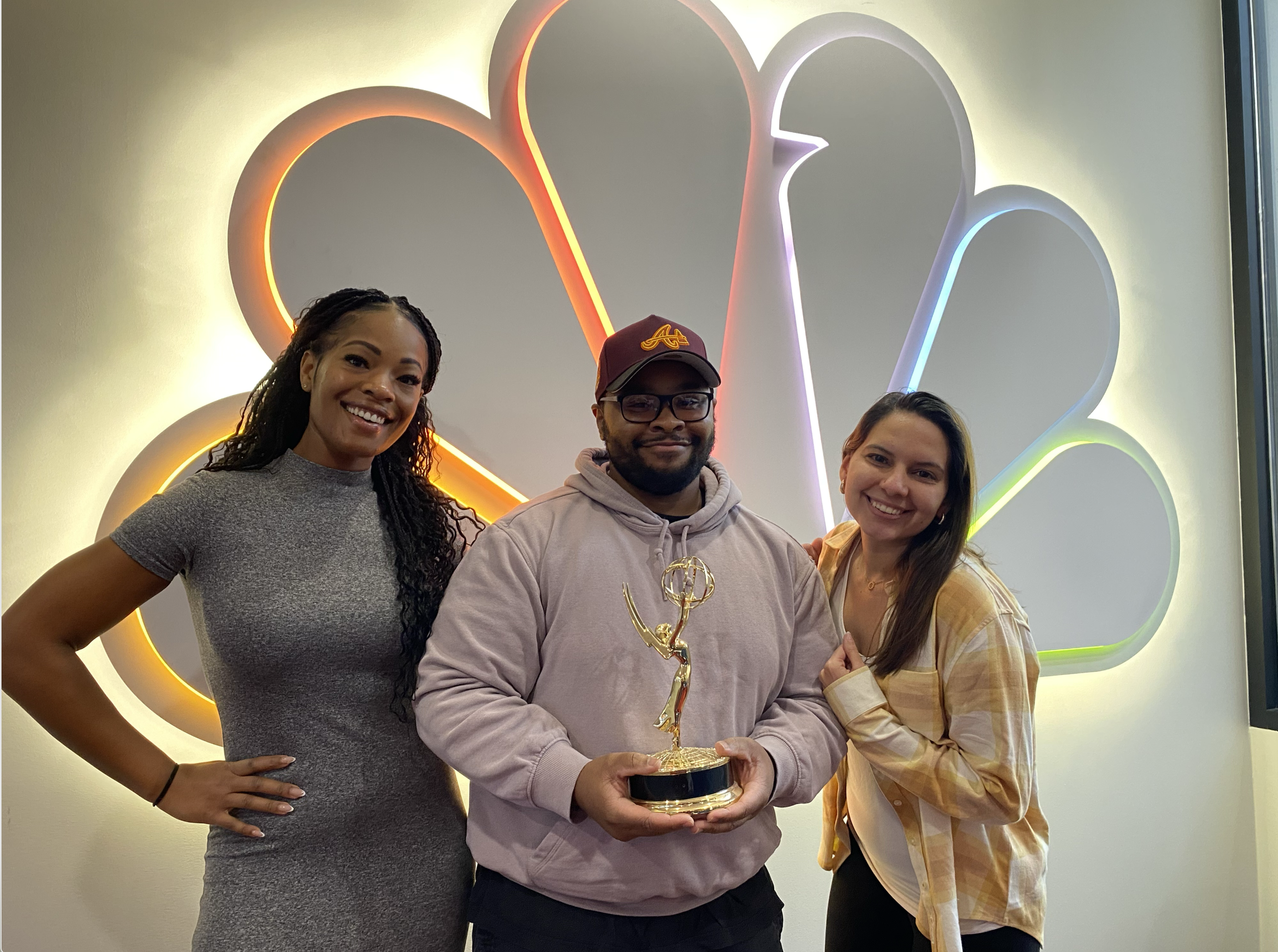 Three smiling people standing in front of a neon NBC peacock logo, with the person in the middle holding an Emmy award.
