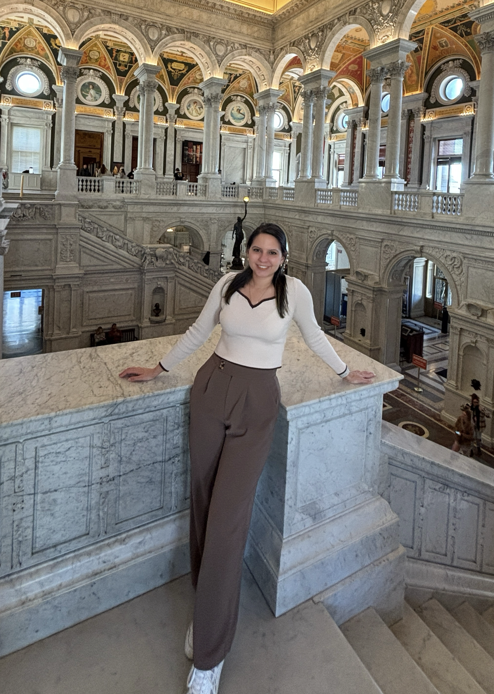 A woman standing on a marble staircase inside a grand historical building with ornate architecture, arches, and columns, smiling at the camera.