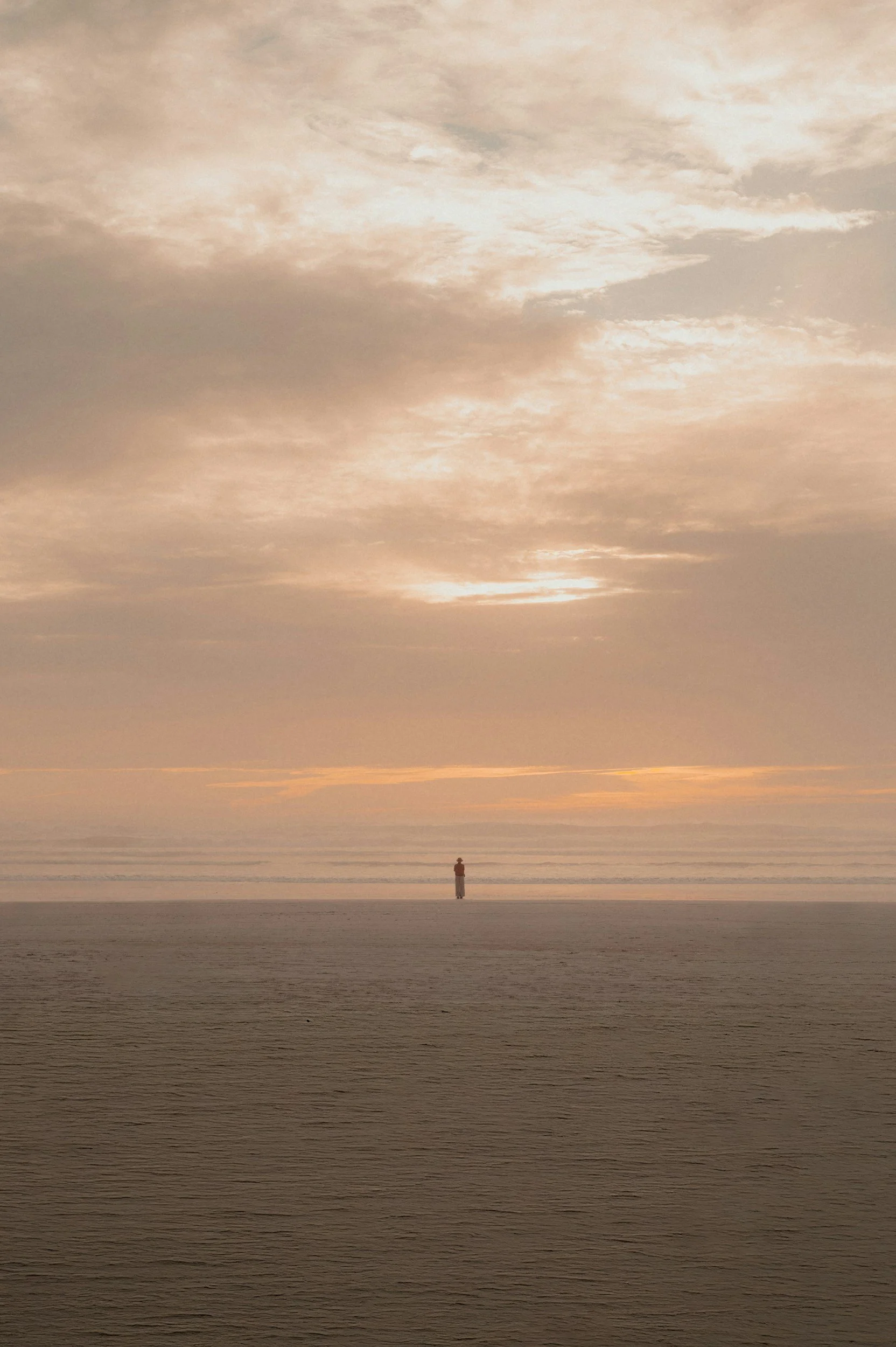 A person standing alone on a beach at sunset, with a cloudy sky and calm ocean waves in the background.